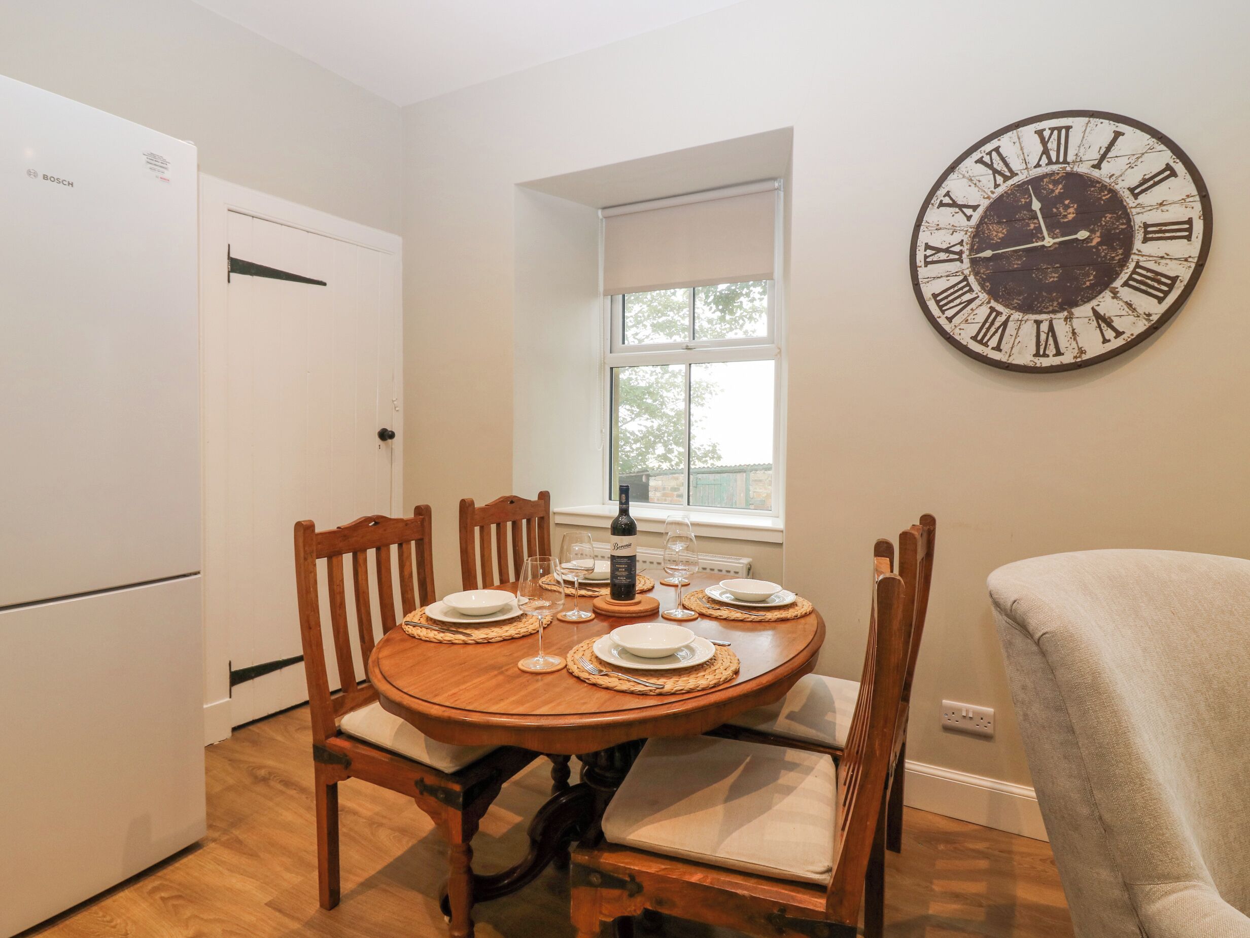 A dining area with a round wooden table set for four, surrounded by wooden chairs with cushioned seats. A large wall clock with Roman numerals is mounted above the table.