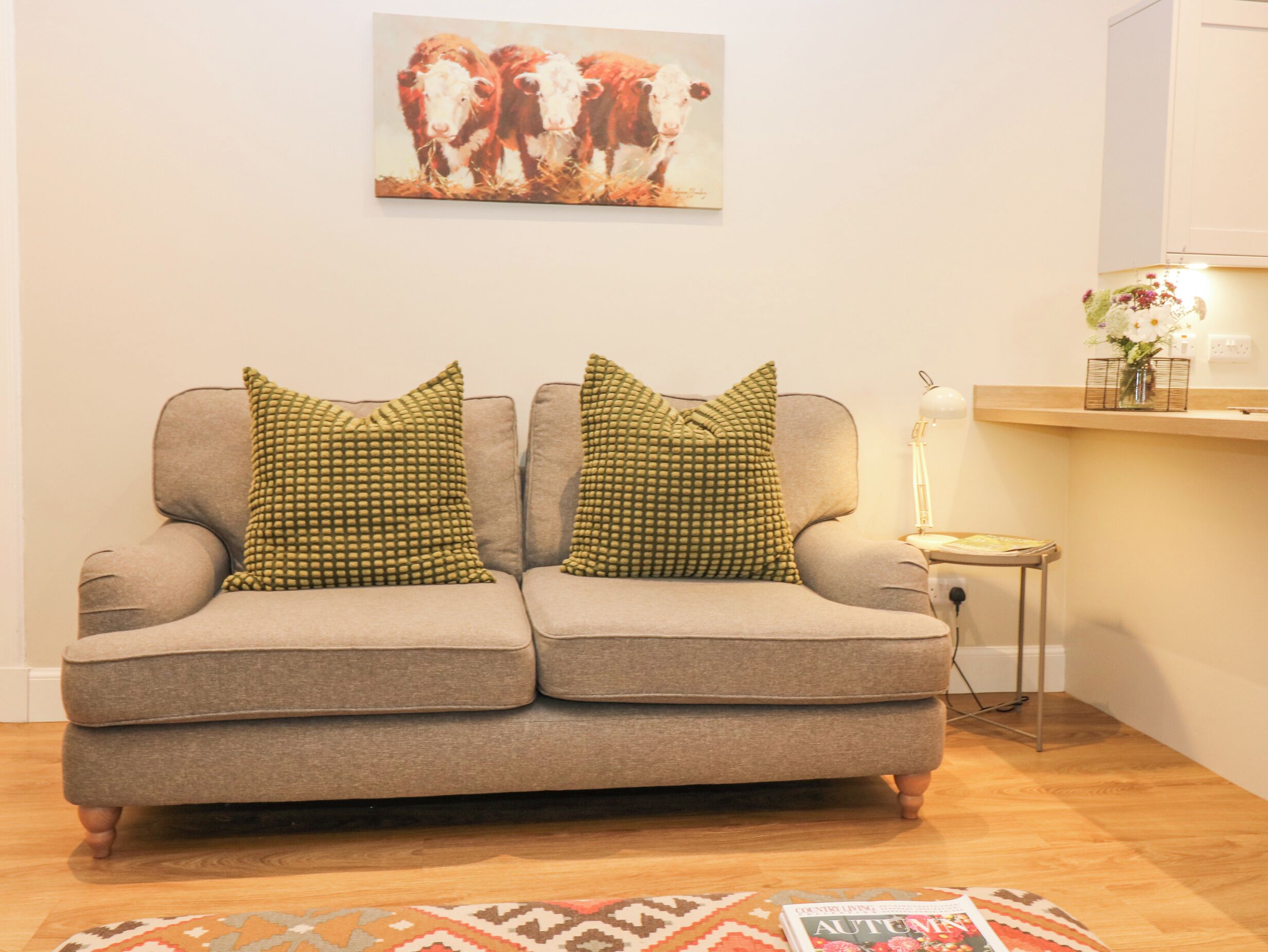 A living room with a grey sofa, two patterned cushions, a painting of cows, a small table with a lamp, and a magazine on the floor.