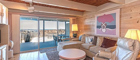 A spacious living room with a wooden ceiling, a large sliding glass door leading to an outdoor area, a beige sofa, a leather armchair, and a round coffee table.