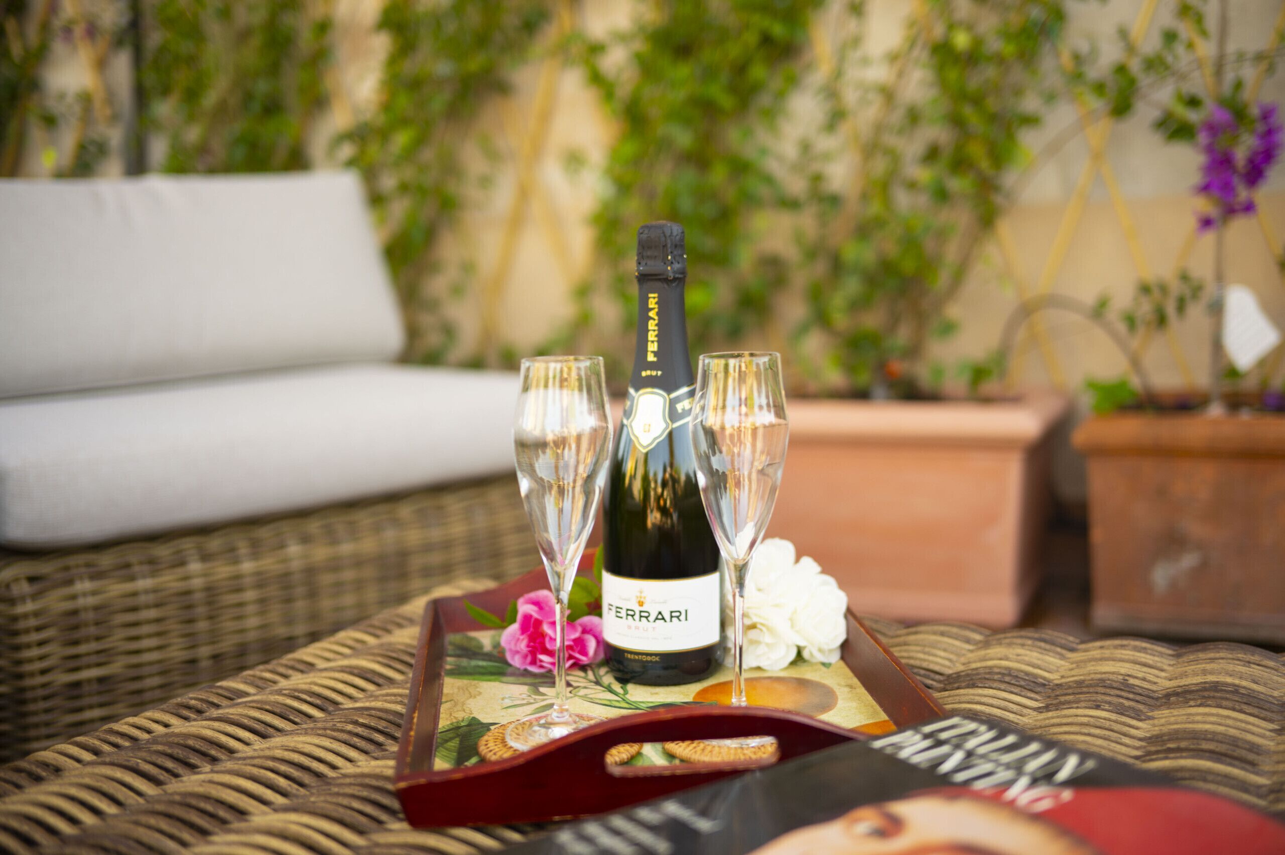A bottle of Ferrari wine, two glasses, and a tray with a floral design on a wicker sofa.
