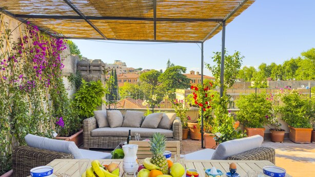 A covered outdoor seating area with a wicker sofa, a table set with fruits and drinks, and potted plants. - Trastevere Outstanding  4 BR Terraced Apartment (Roma)