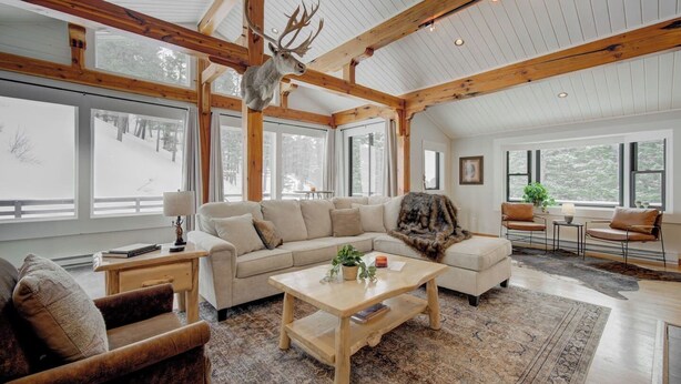 Un salon spacieux avec un grand canapé, une table basse et une cheminée. De grandes fenêtres et un plafond en bois complètent l’ensemble.