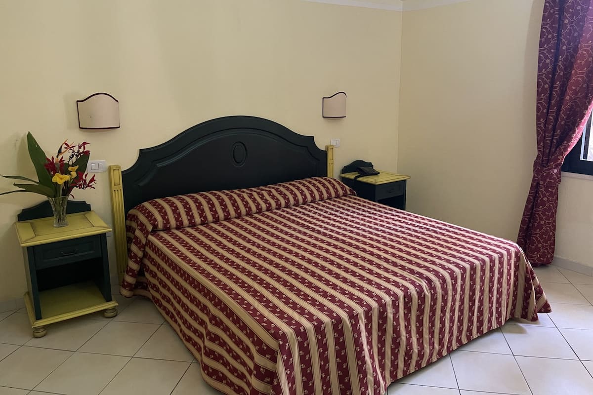 A bedroom with a large bed, two bedside tables, a nightstand, a wall-mounted lamp, and a window with curtains.
