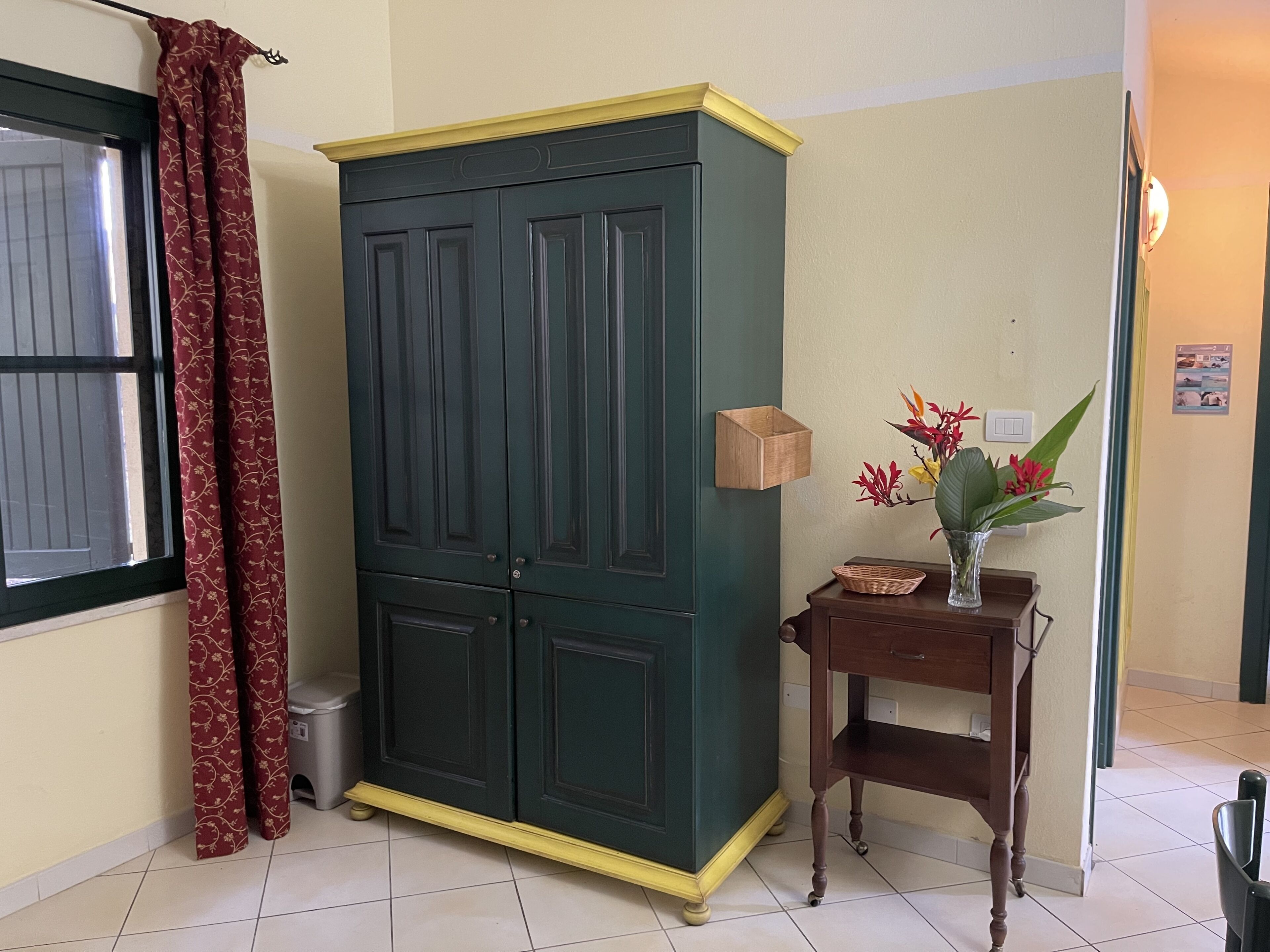 A room with a green cabinet, a small wooden table with a vase of flowers, and a window with red curtains.