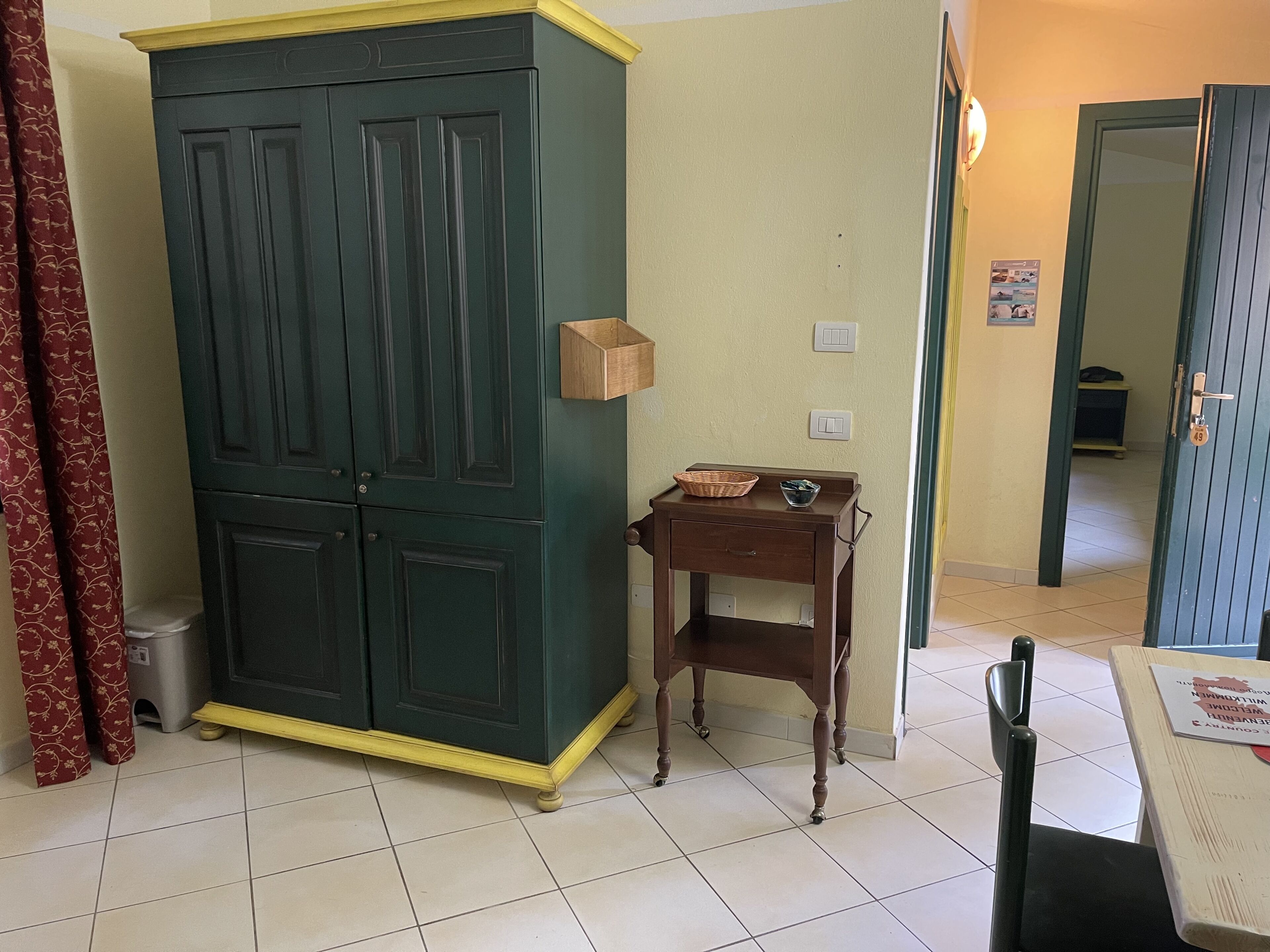 A room with a large green wardrobe, a small wooden table with a basket and bowl, and a tiled floor.