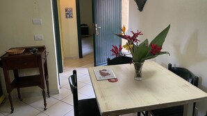 A dining area with a table, chairs, a small side table with a basket and bowl, a vase with flowers, and a door leading to another room. - Ala Birdi Residence  (Arborea)
