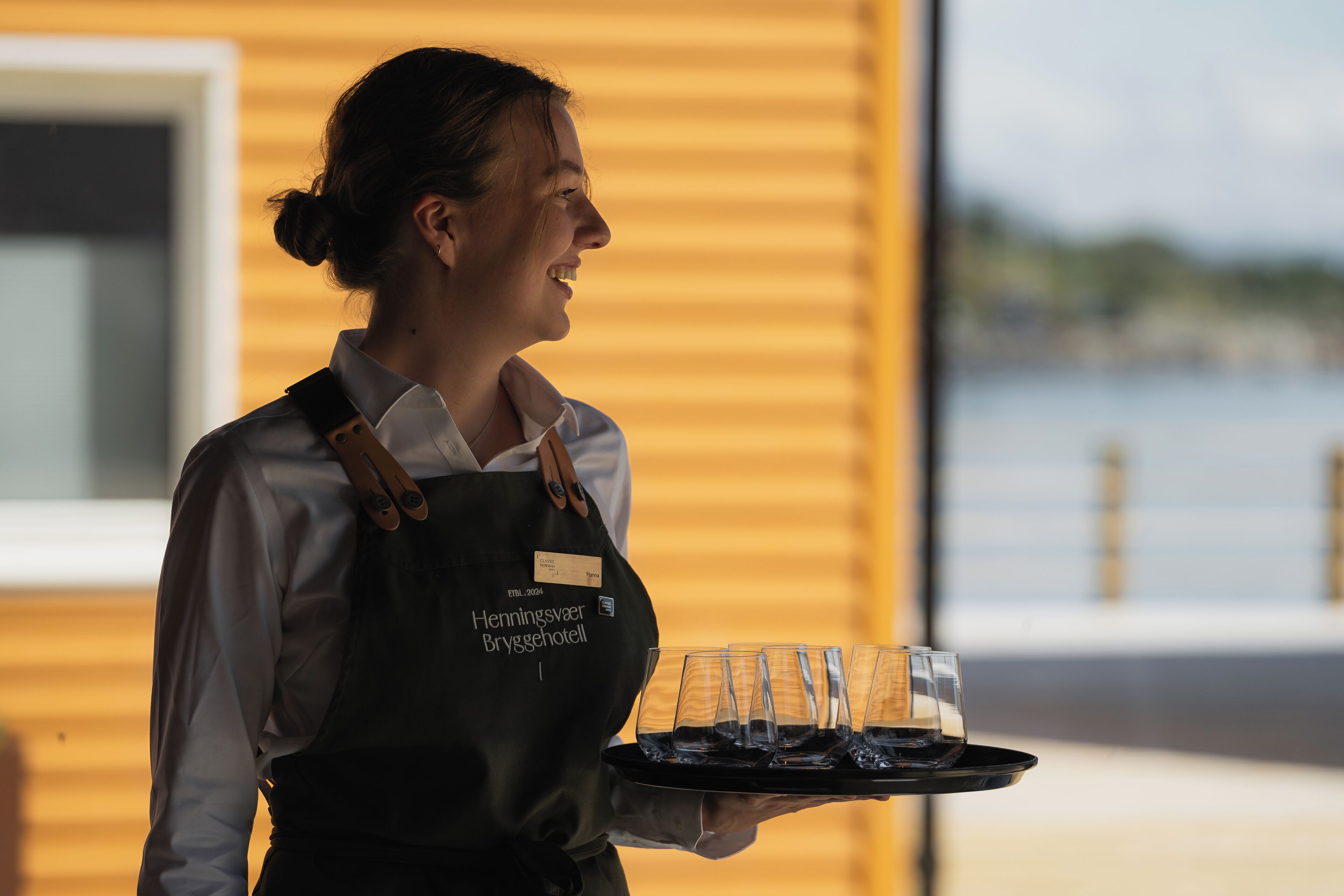 A person holding a tray with glasses and a bottle, wearing an apron with a name tag.