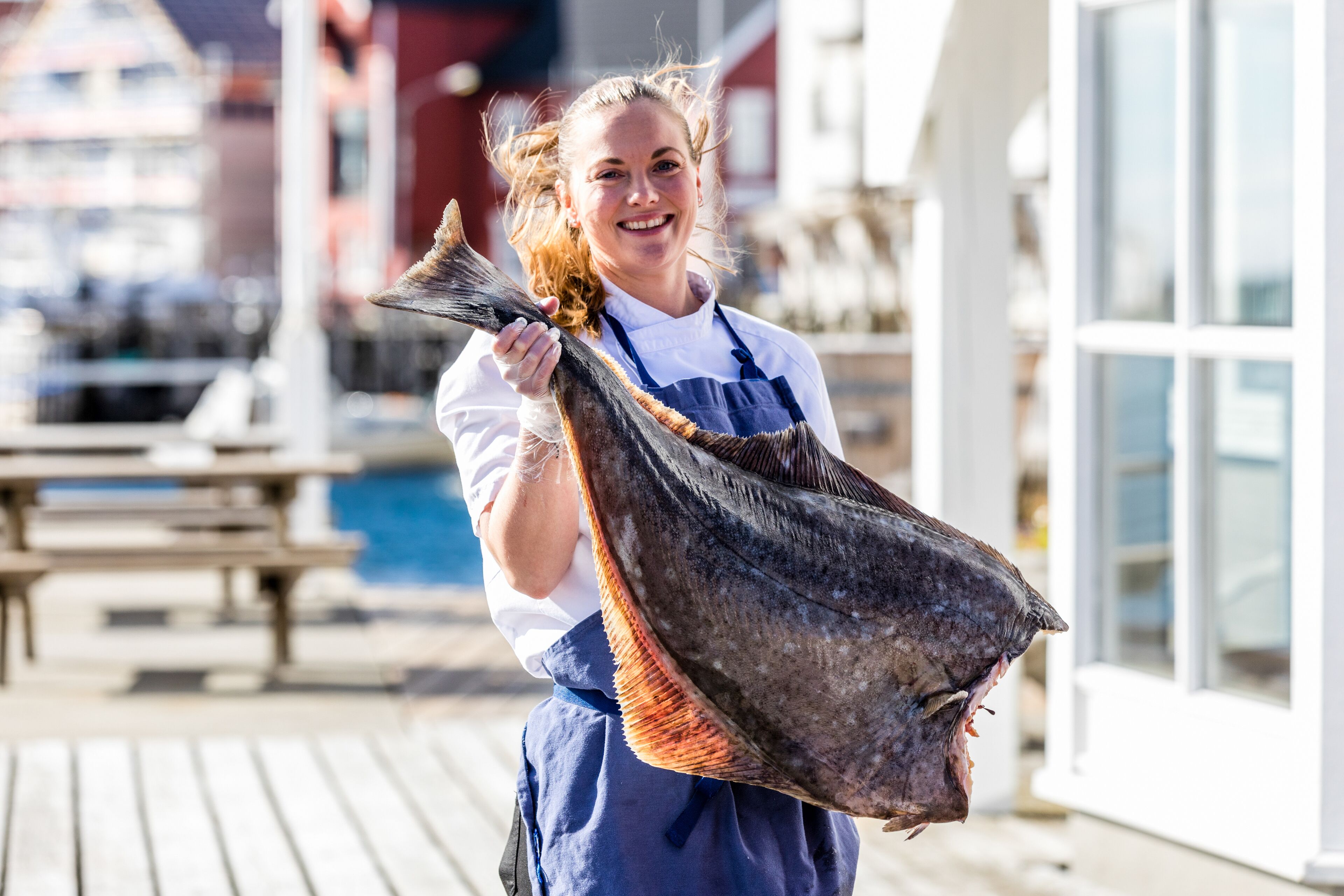 A person holding a large fish, possibly a sea bass, outdoors with a wooden deck and buildings in the background.