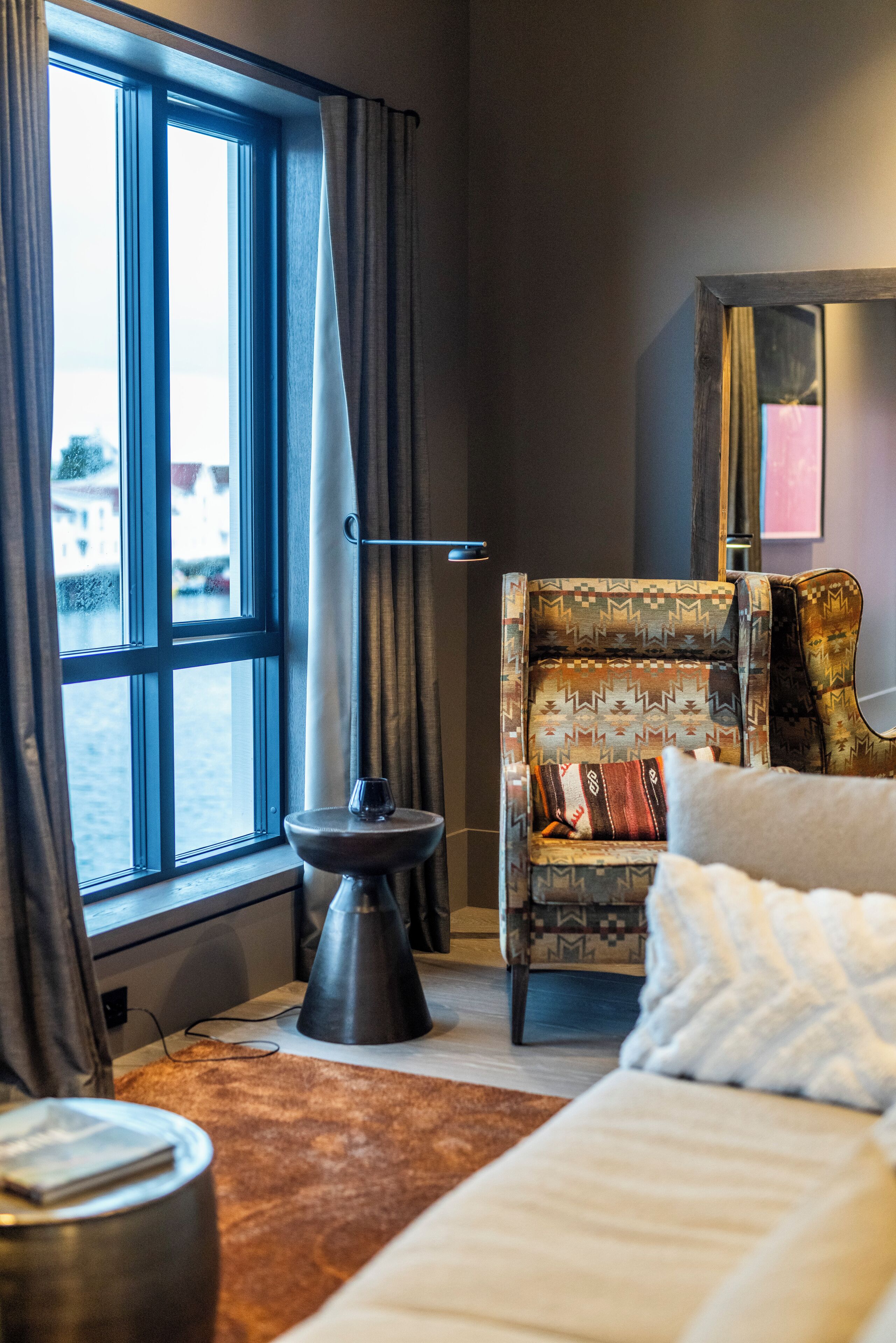 Ein Zimmer mit einem großen Fenster, einem gemusterten Sessel, einem kleinen Tisch mit einem Glas und einem weißen Sofa.