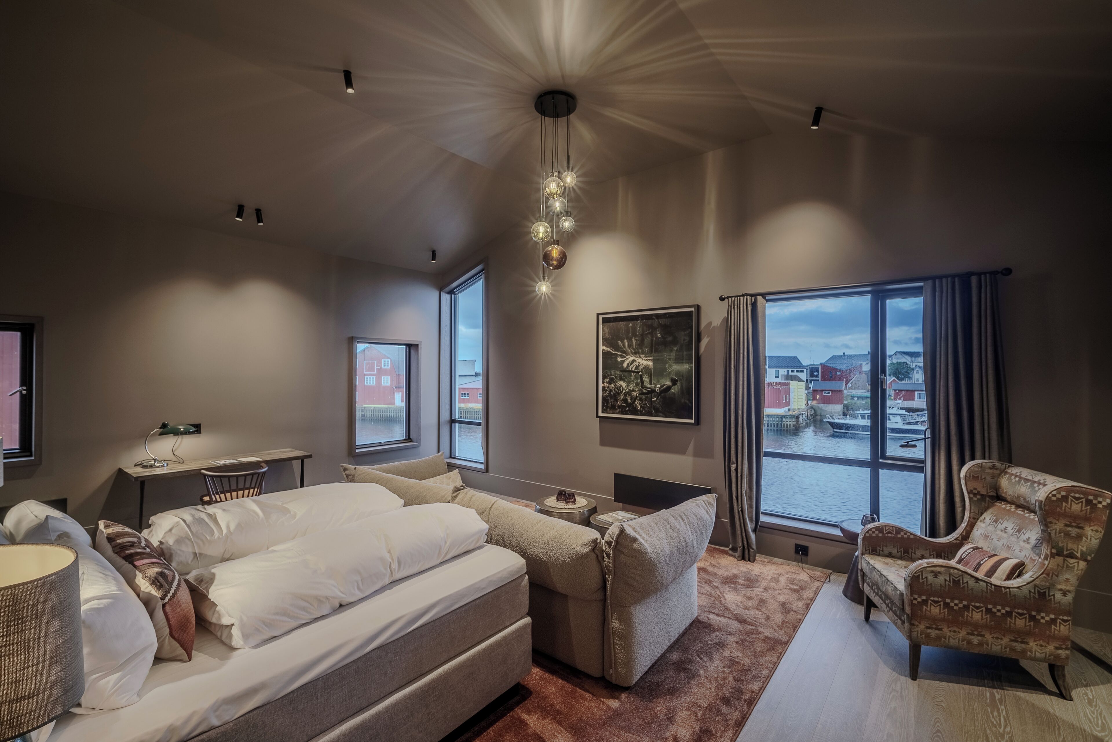 A modern hotel room with a large bed, a sofa, a chair, and a view of a cityscape through large windows.