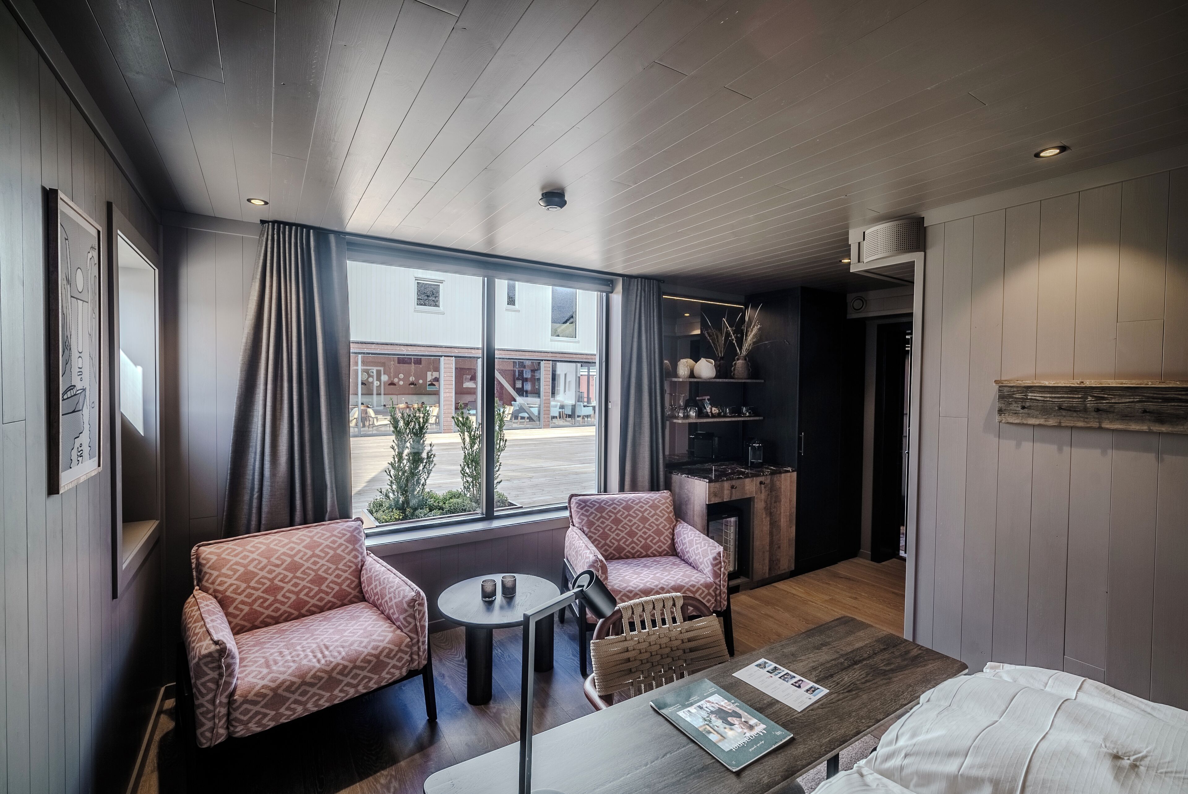 A modern room with two patterned armchairs, a small round table, and a wooden coffee table. A large window offers a view of the outside.