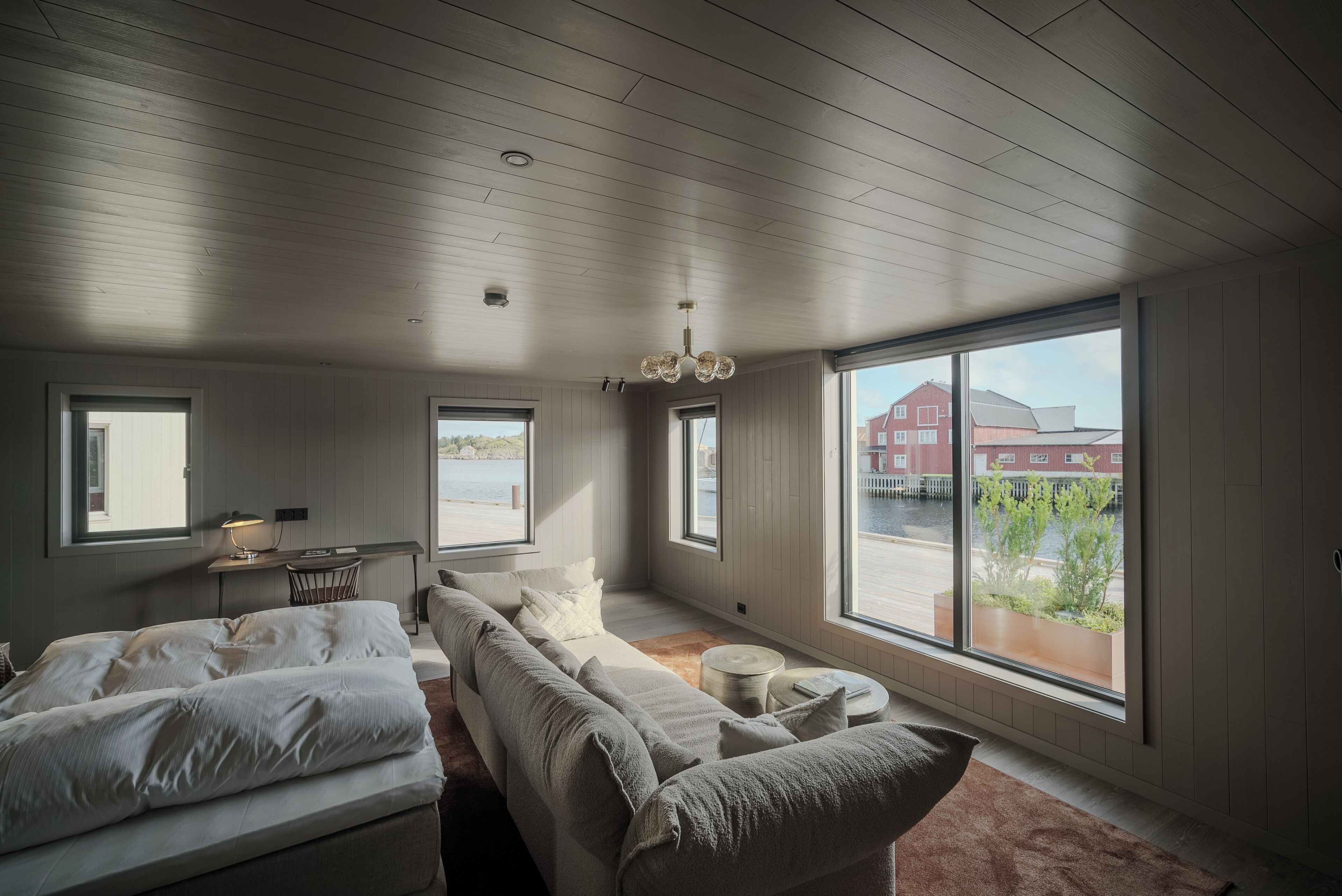 A bedroom with a large bed, a sofa, a small table, and a window with a view of buildings and greenery.