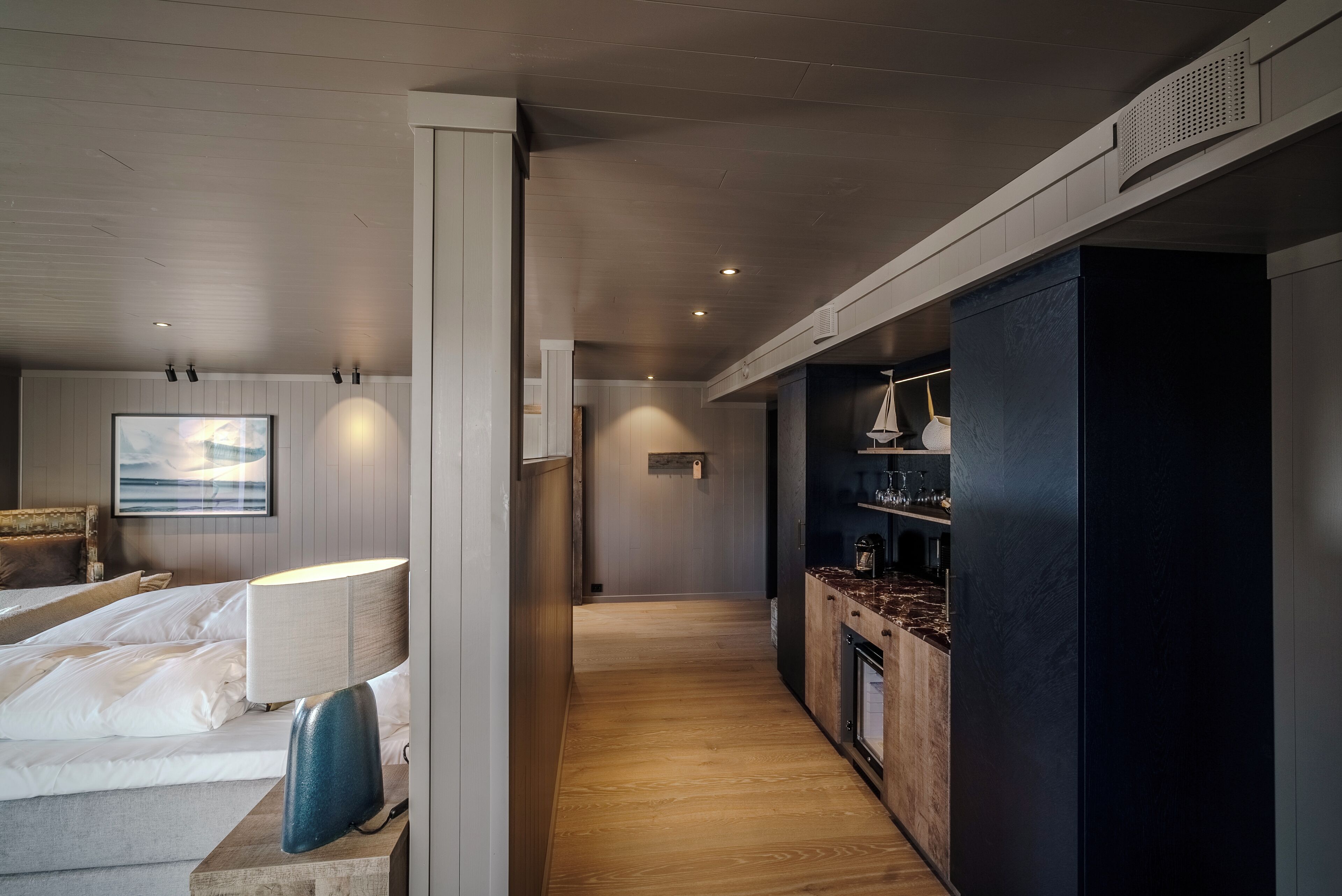 A modern hotel room with a wooden floor, a bed with white linens, a nightstand, a desk with a lamp, a dark-colored wardrobe, and a small kitchen area with a marble countertop and a coffee machine.
