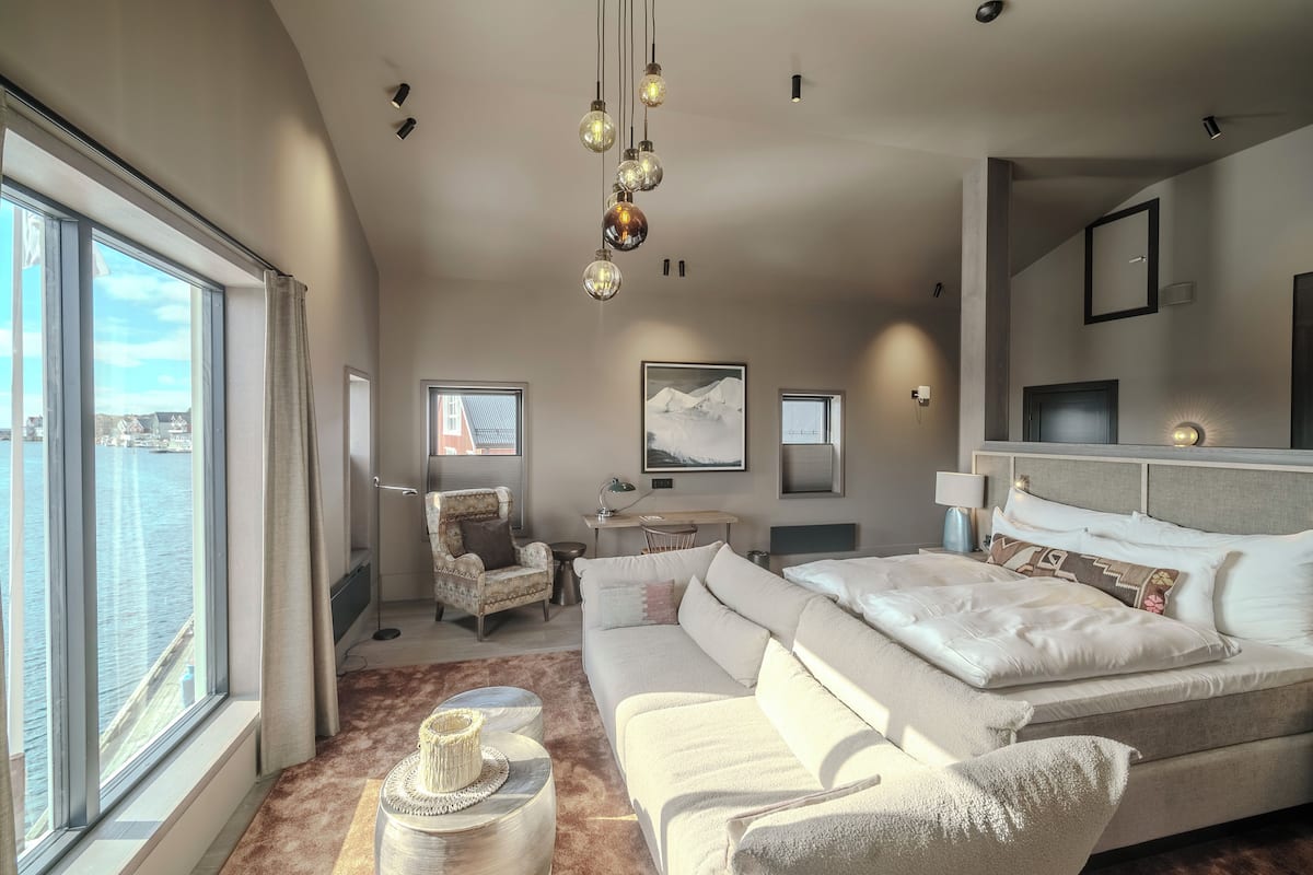 Une chambre d’hôtel moderne, dotée d’un grand lit, d’un canapé, d’un bureau et d’une chaise. Une grande fenêtre offre une vue sur l’eau.