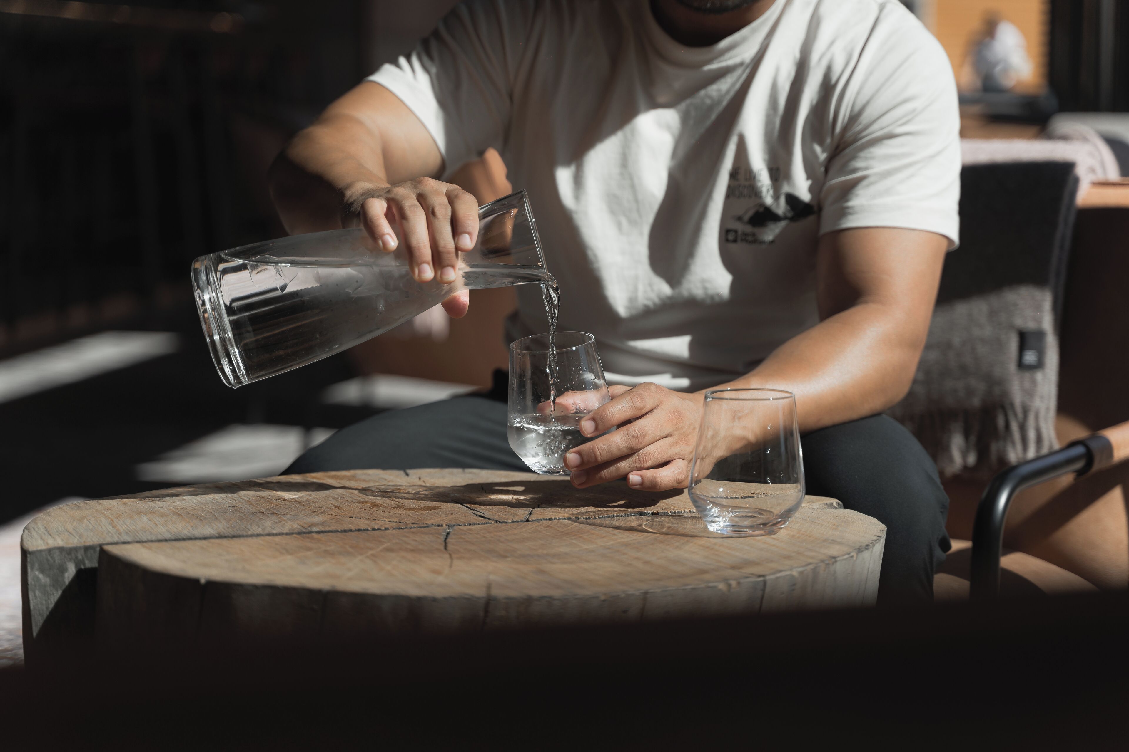 A person pouring water from a clear glass pitcher into a glass on a wooden table.