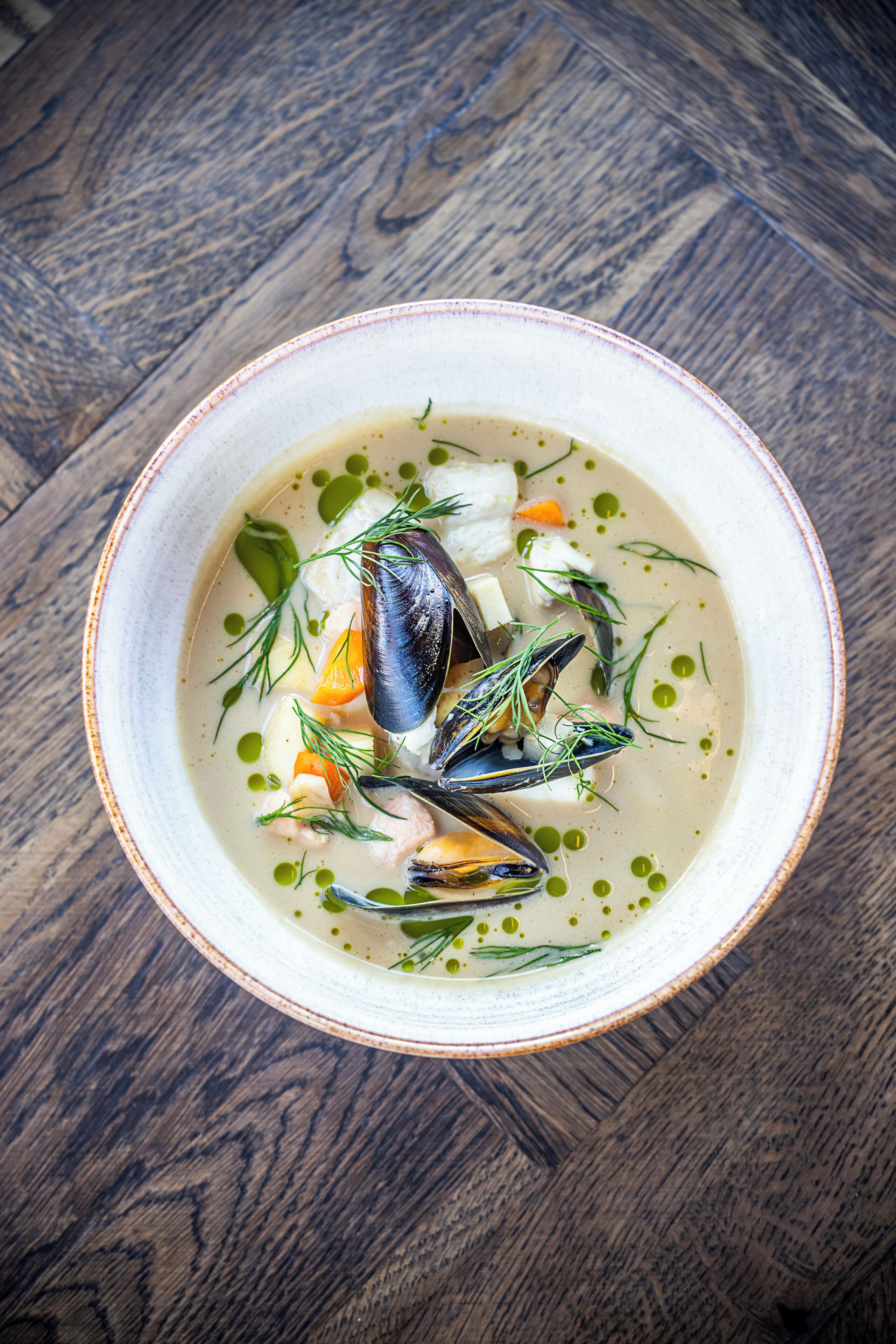 A bowl of seafood soup with mussels, carrots, and herbs on a wooden table.