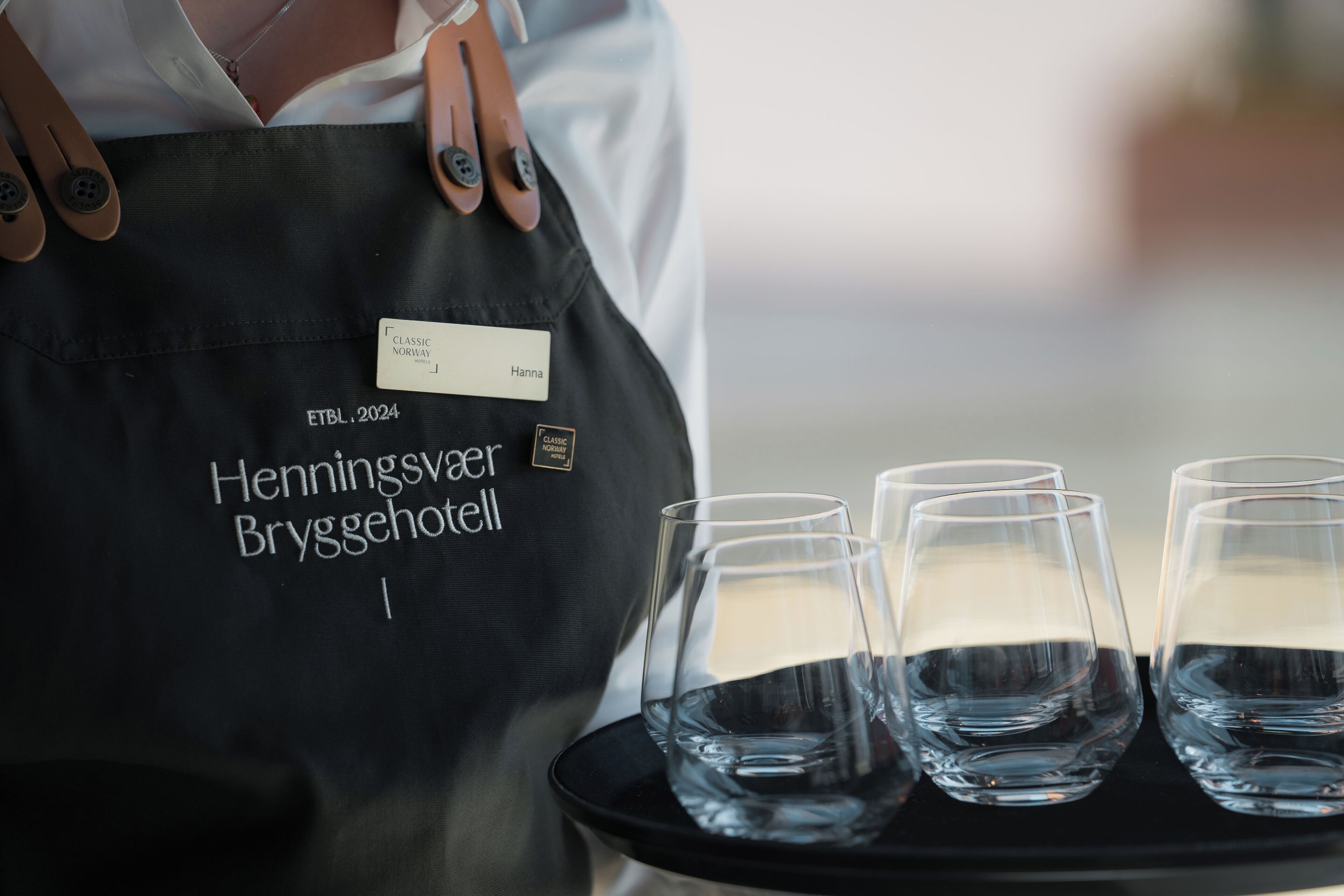 A person wearing a black apron with a name tag and the text "Henningsvær Bryggehotel" on it, holding a tray with four empty glasses of water.