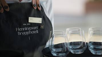 A person wearing a black apron with a name tag and the text "Henningsvær Bryggehotel" on it, holding a tray with four empty glasses of water.