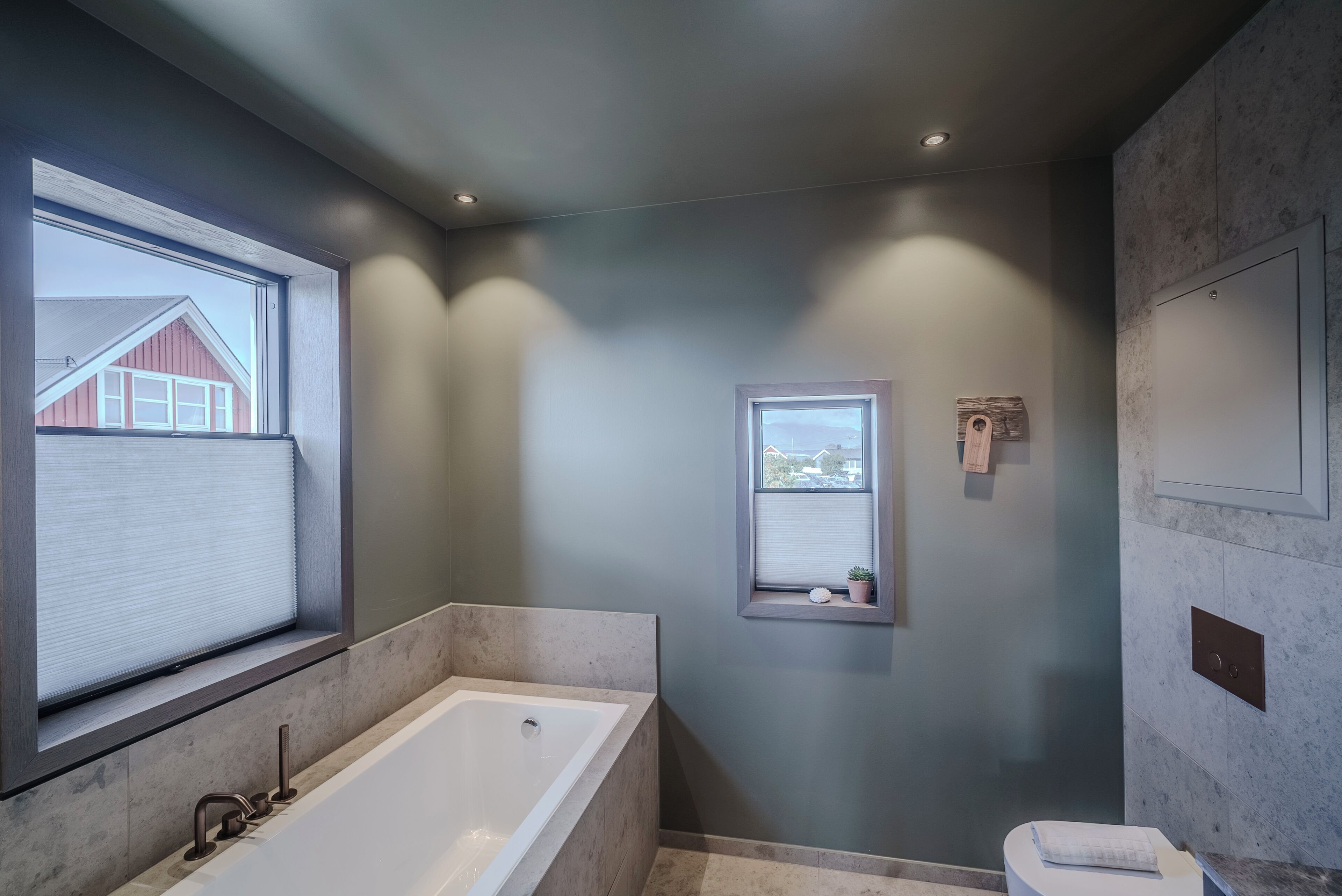 Un baño moderno con una bañera grande, un ventanal con vistas a un edificio rojo y un ventanuilla con vistas al paisaje urbano.