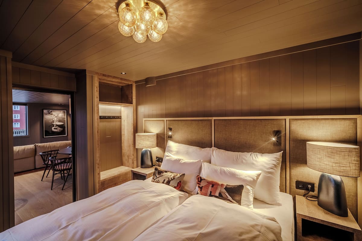 Une chambre d’hôtel avec un grand lit, une tête de lit en bois, deux tables de chevet avec des lampes, une petite table avec des chaises et un tableau encadré au mur.