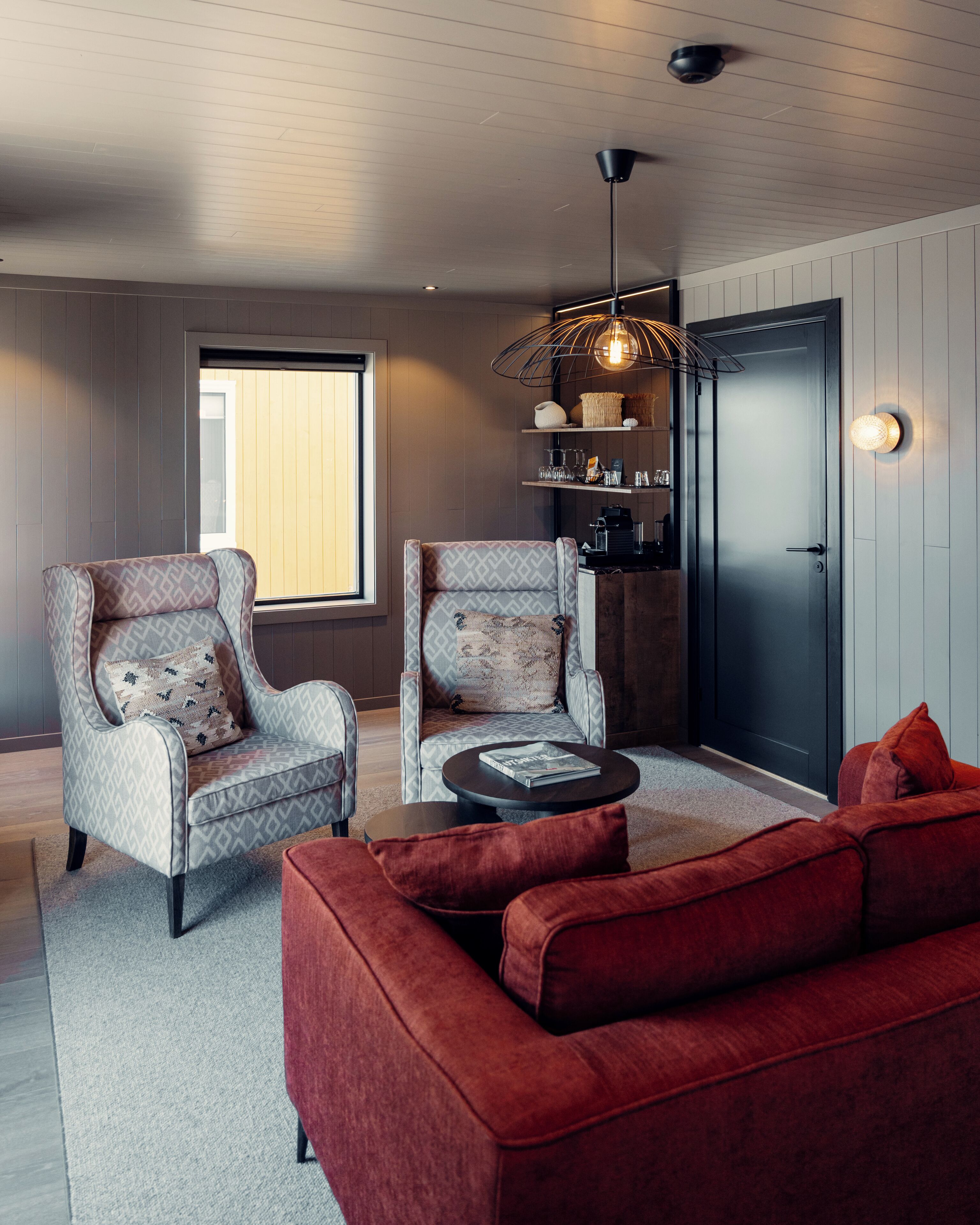 Un salon moderne avec deux fauteuils à motifs, un canapé rouge, une table basse ronde, une porte noire et une fenêtre avec des stores.
