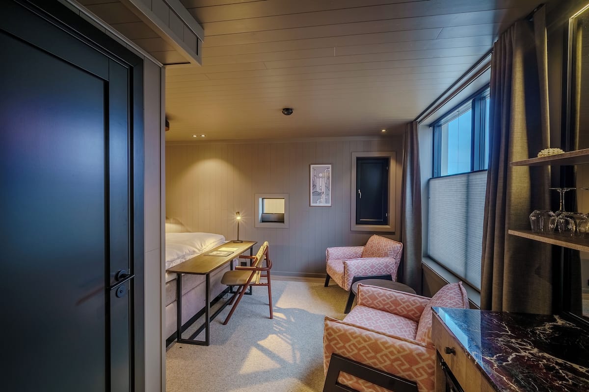 Une chambre d’hôtel de taille réduite, comprenant un lit, un bureau, deux chaises, une petite table et une fenêtre avec des rideaux.