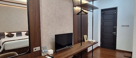 A modern hotel room with a wooden floor, a large flat-screen TV, a mirror, and a bench with a towel and a small table.