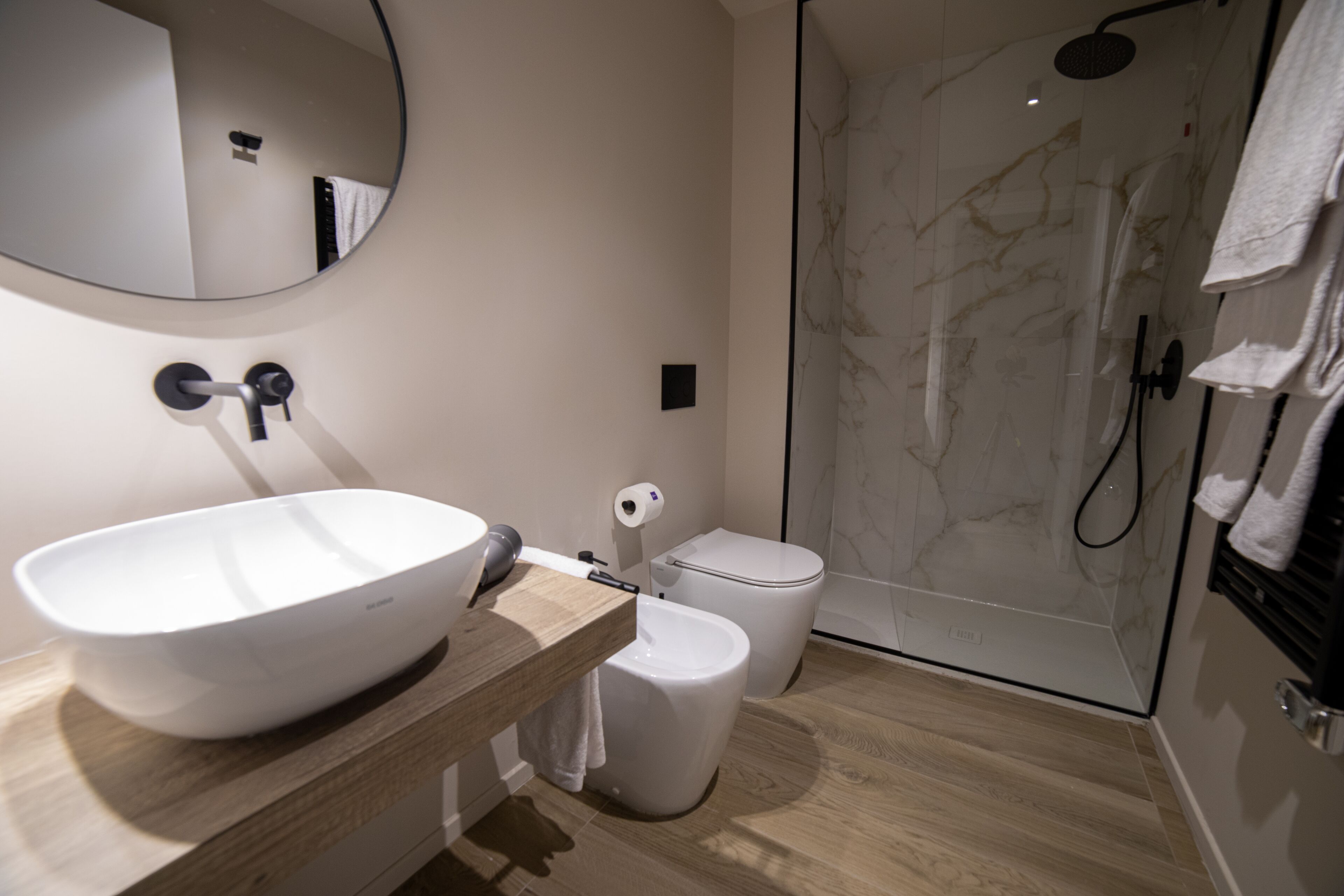 Un baño moderno con un espejo redondo grande, un lavabo, un inodoro y una ducha a ras de suelo.
