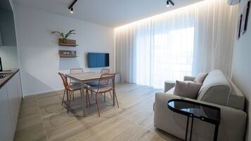 Uma sala de estar moderna com área de jantar, sofá, televisão e cozinha ao fundo.