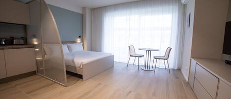 Une chambre d’hôtel moderne comprenant un lit, un coin repas avec deux chaises, une petite kitchenette et une grande fenêtre avec des rideaux.