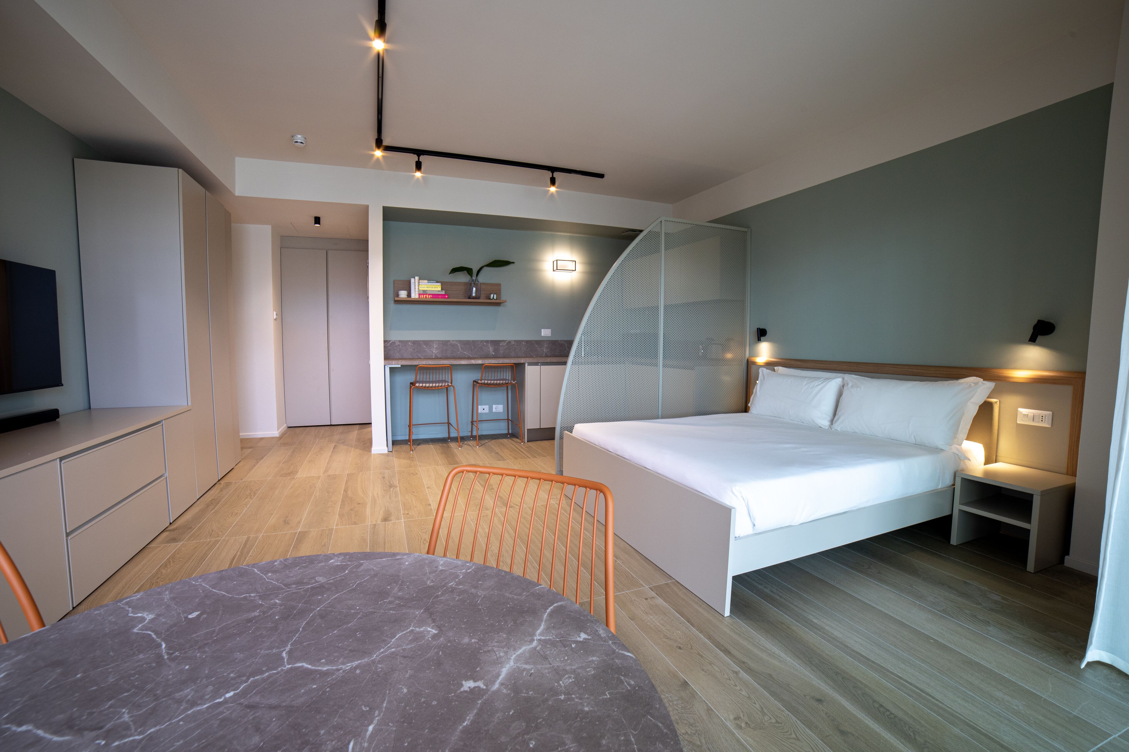 A modern hotel room with a bed, a desk, a chair, a TV, and a small kitchen area.