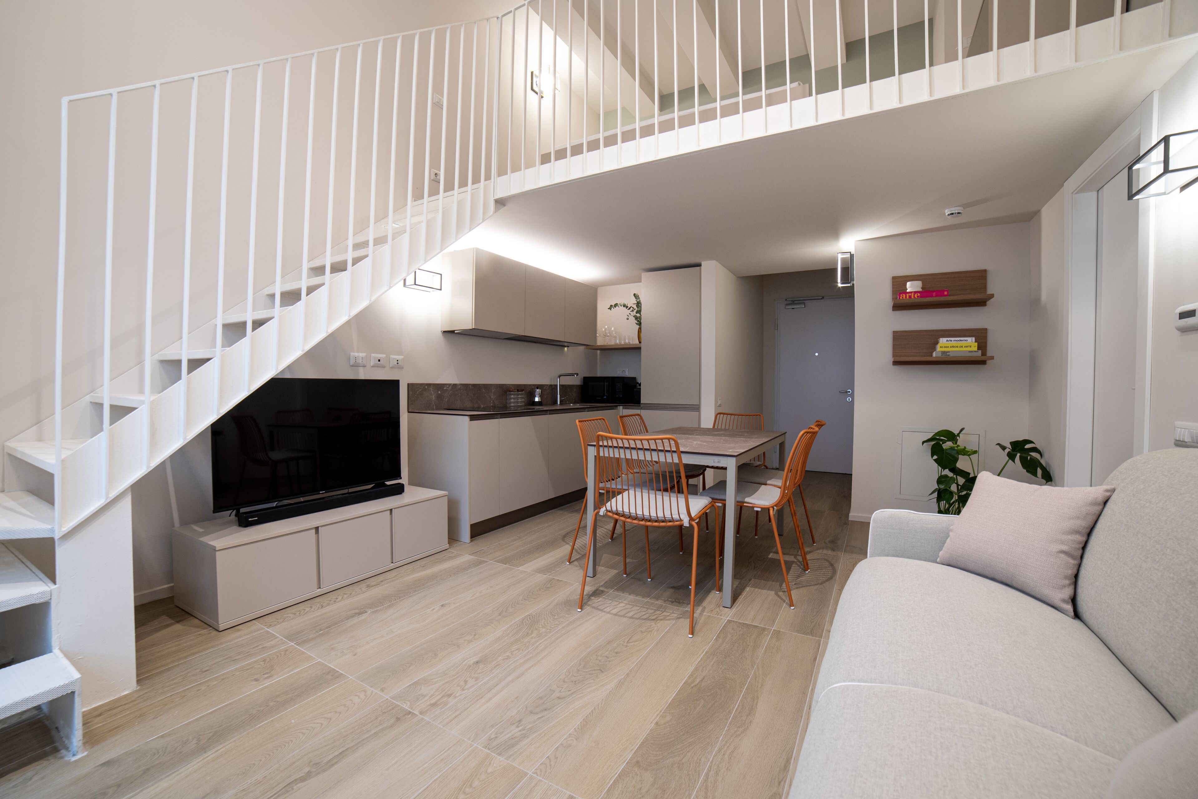 A modern apartment with a staircase leading to a loft, a dining area with a table and chairs, a sofa, and a TV in the living room.