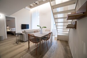 Egy modern, nyitott tér, amelyben étkező, kanapé, a falra szerelt televízió és egy felső szintre vezető lépcső található.