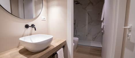 Une salle de bain moderne avec un lavabo sur colonne blanc, un meuble-lavabo en bois, un miroir rond et une douche aux murs en marbre.