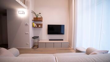 Un soggiorno moderno con una televisione a schermo piatto montata su una parete bianca, un piccolo tavolino e una libreria a parete con libri e una pianta.