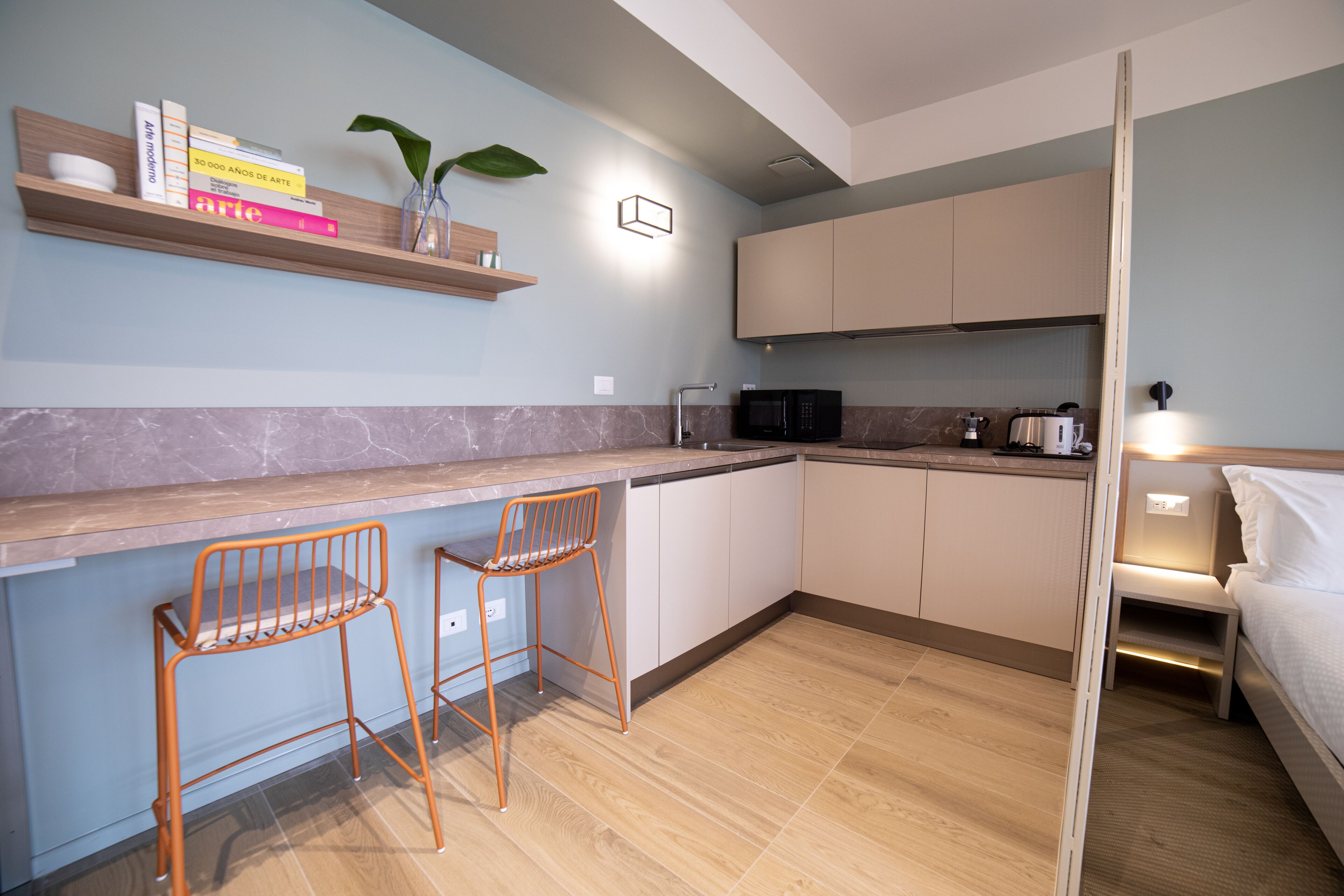 A compact kitchen with a wooden countertop, two orange chairs, a microwave, and a bed with a nightstand.