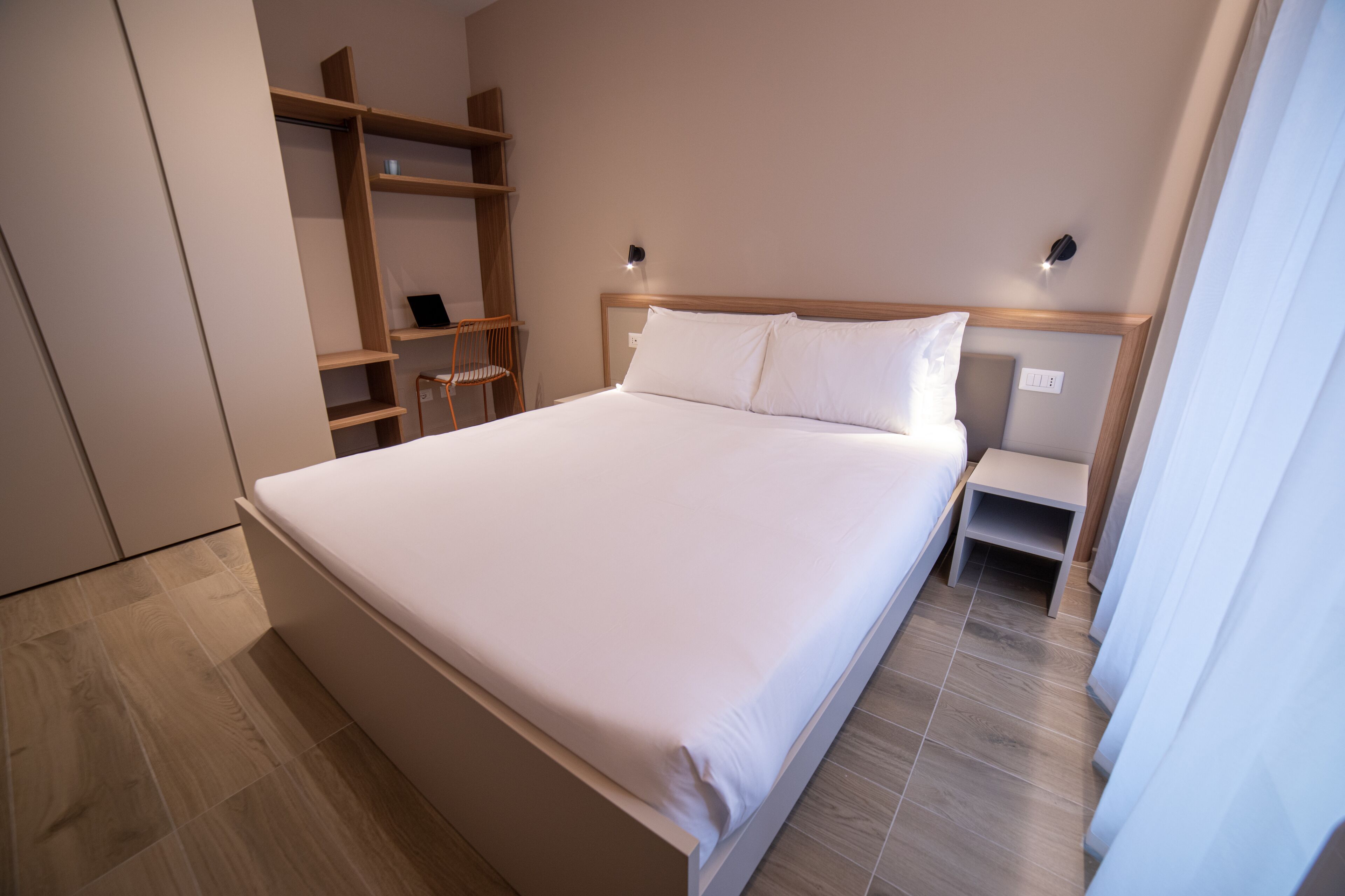 A neatly arranged bedroom with a large bed, a wooden wardrobe, a small table, and a chair.
