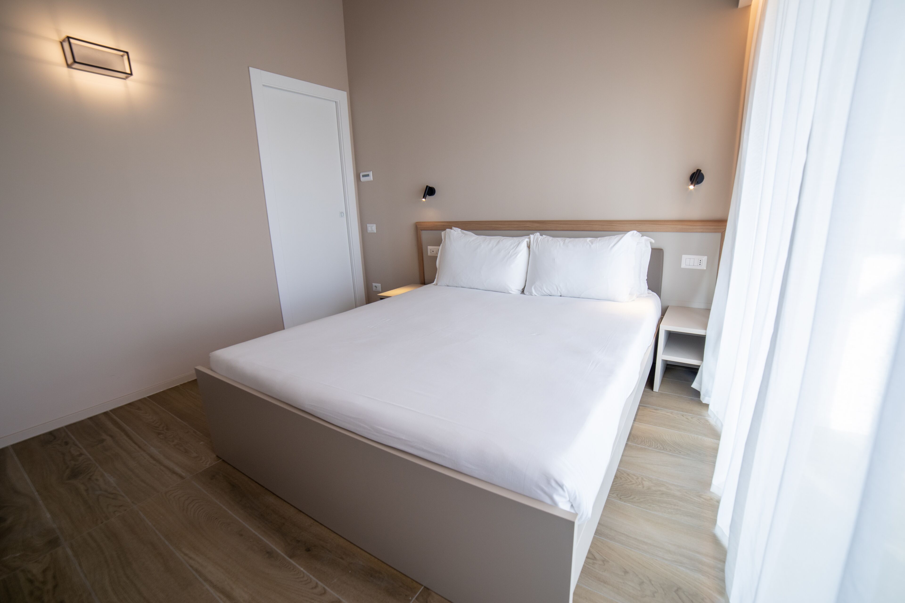 A modern hotel room with a large bed, a bedside table, a nightstand, and a wall-mounted light fixture.
