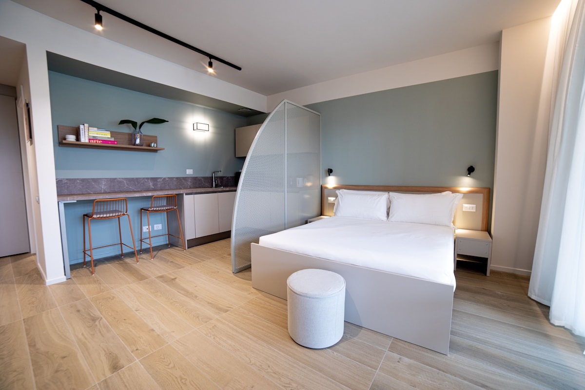 A modern hotel room with a large bed, a small kitchen area with bar stools, and a round ottoman.