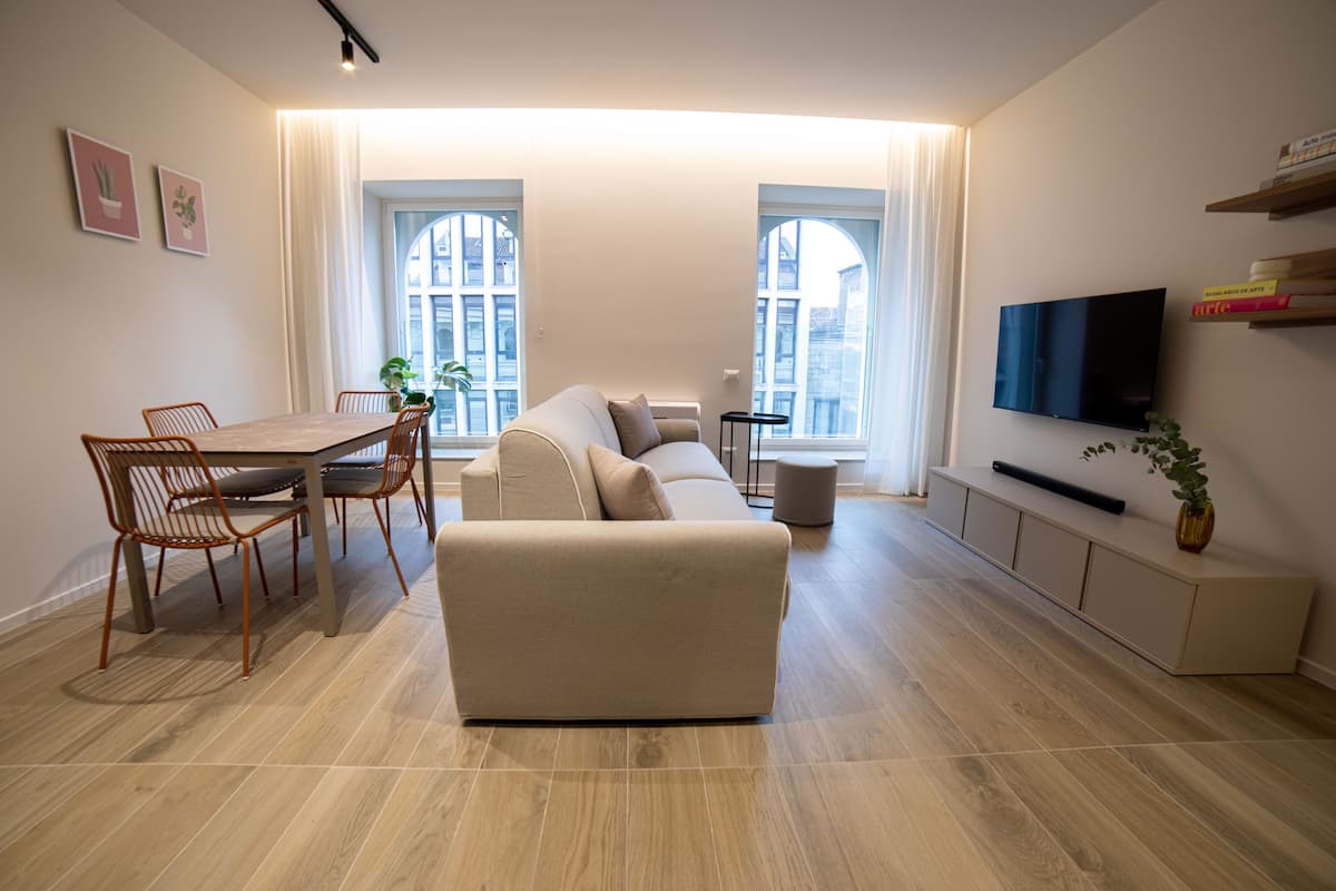 A modern living room with a sofa, dining table, chairs, a TV, and large windows.