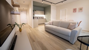 A modern living room with a sofa, a coffee table, and a TV. There is a kitchen area in the background with cabinets and a countertop. - Be Mate Torino Centro (Turin)