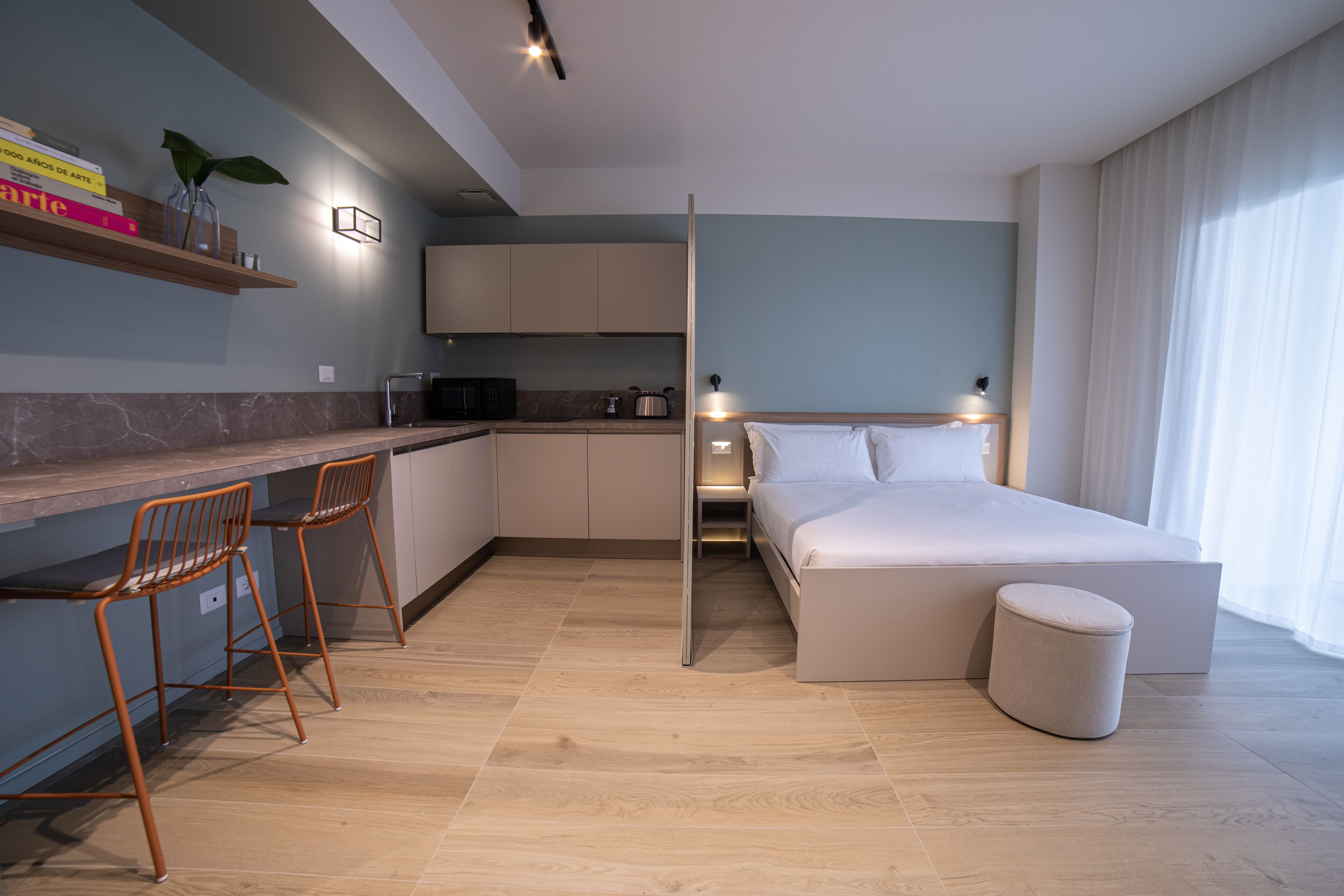 A modern hotel room with a bed, a small kitchenette, a seating area with chairs, and a bedside table.