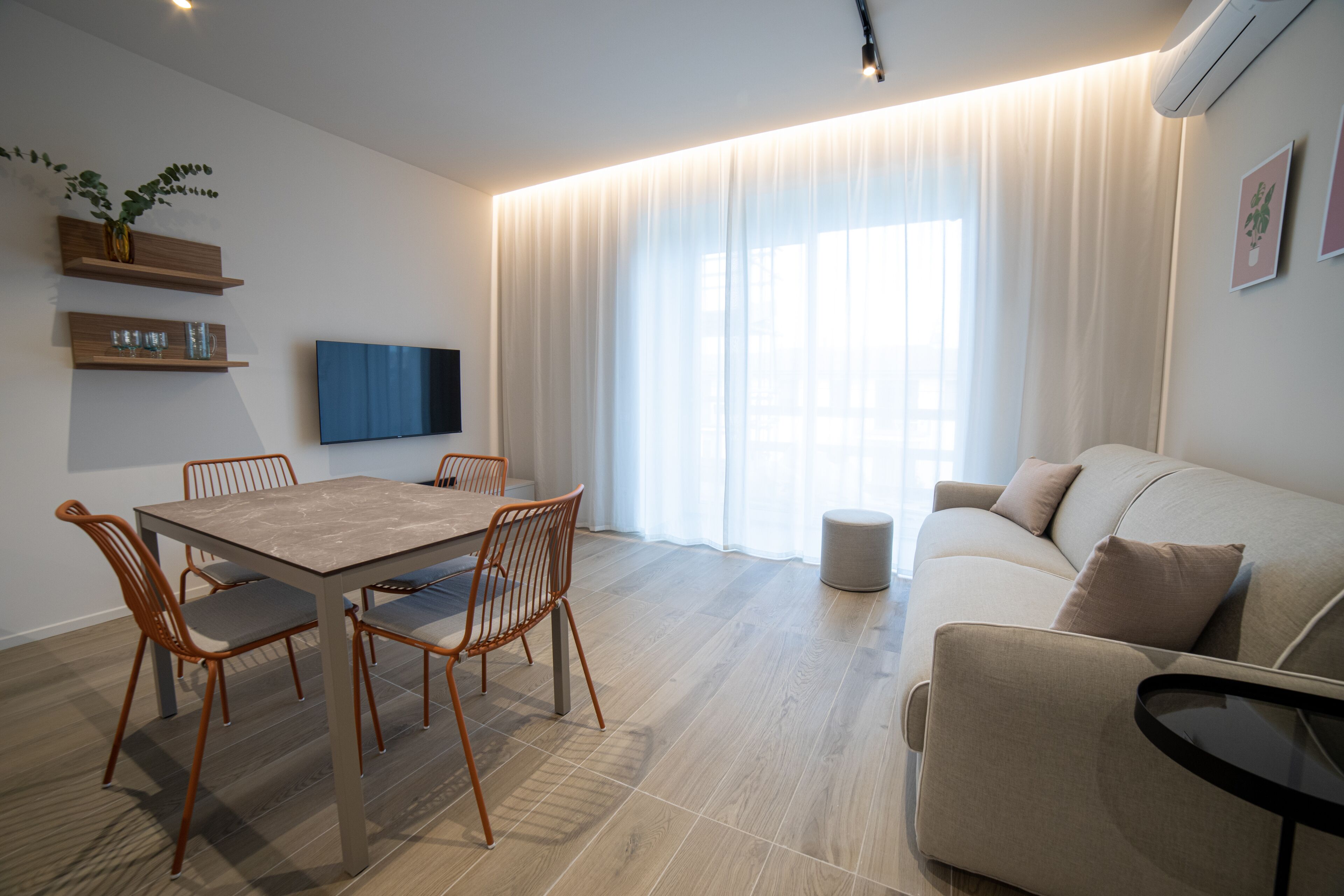 A modern living room with a wooden dining table, four chairs, a sofa, and a TV mounted on the wall.