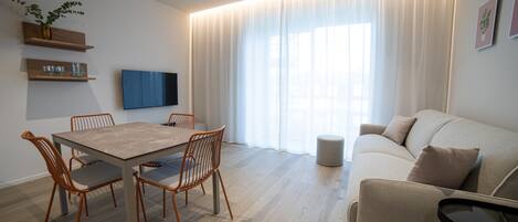 A modern living room with a wooden dining table, four chairs, a sofa, and a TV mounted on the wall.