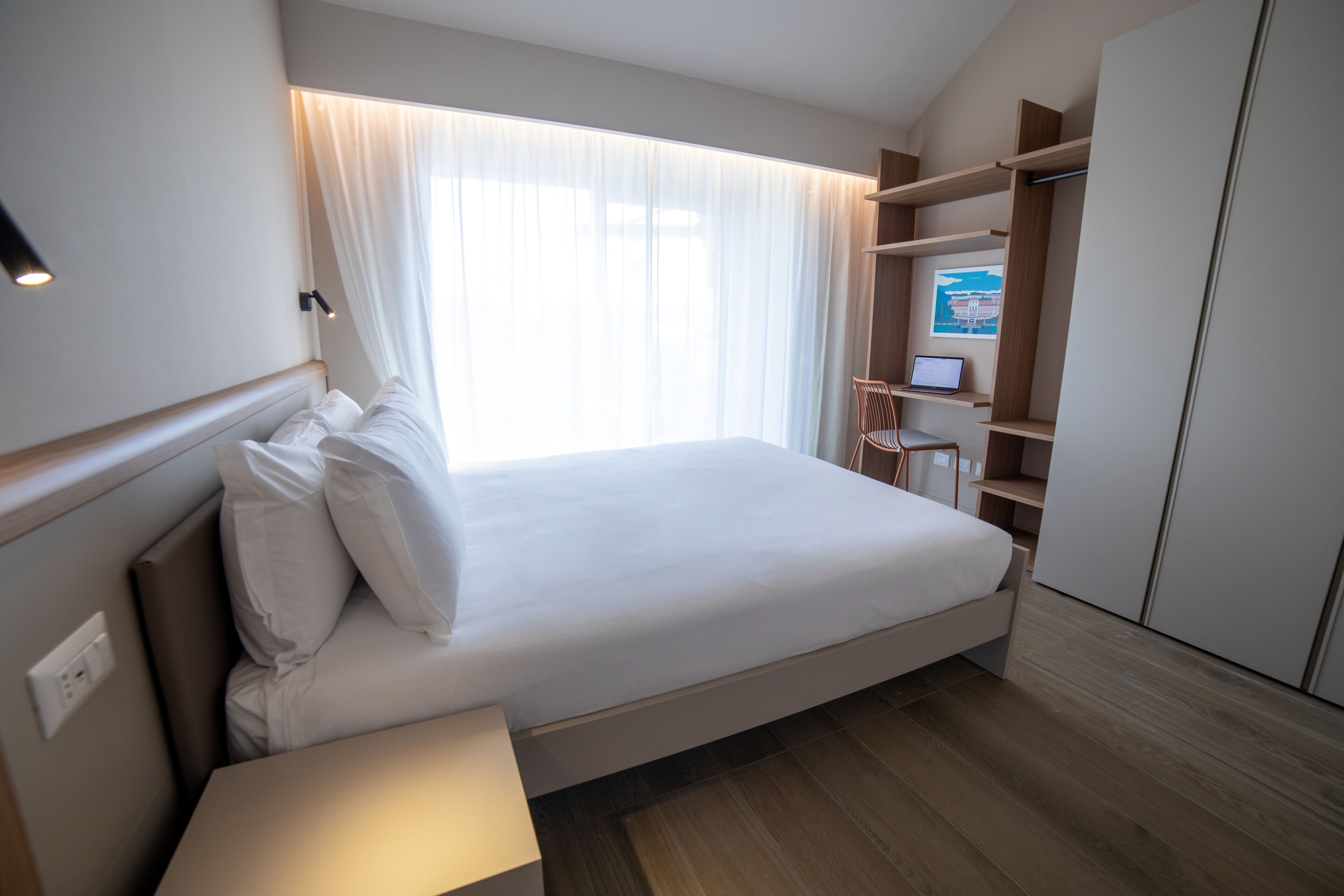 A modern hotel room with a large bed, bedside tables, a desk with a chair, and built-in shelving.