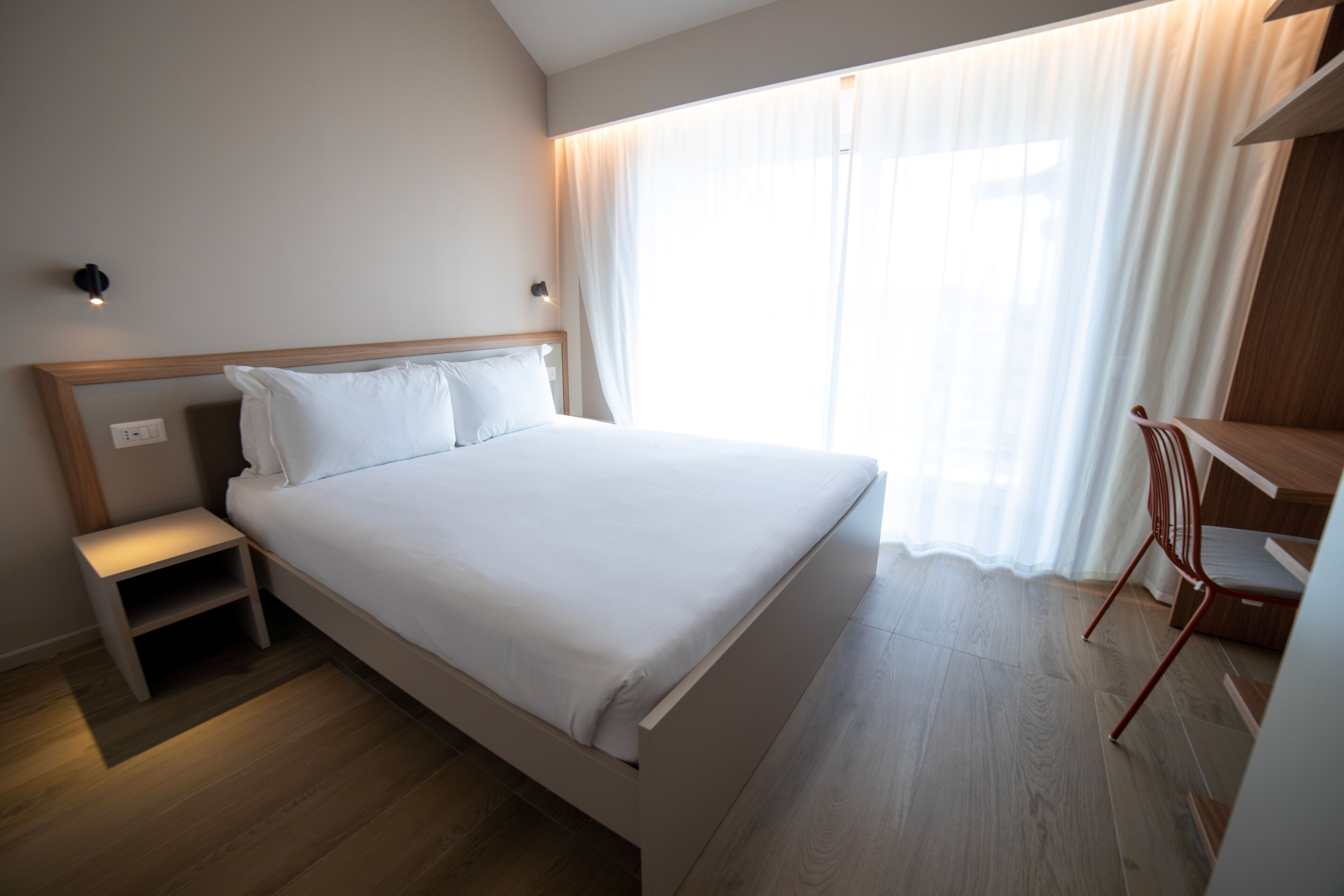 A modern hotel room with a large bed, a bedside table, a desk, and a chair.