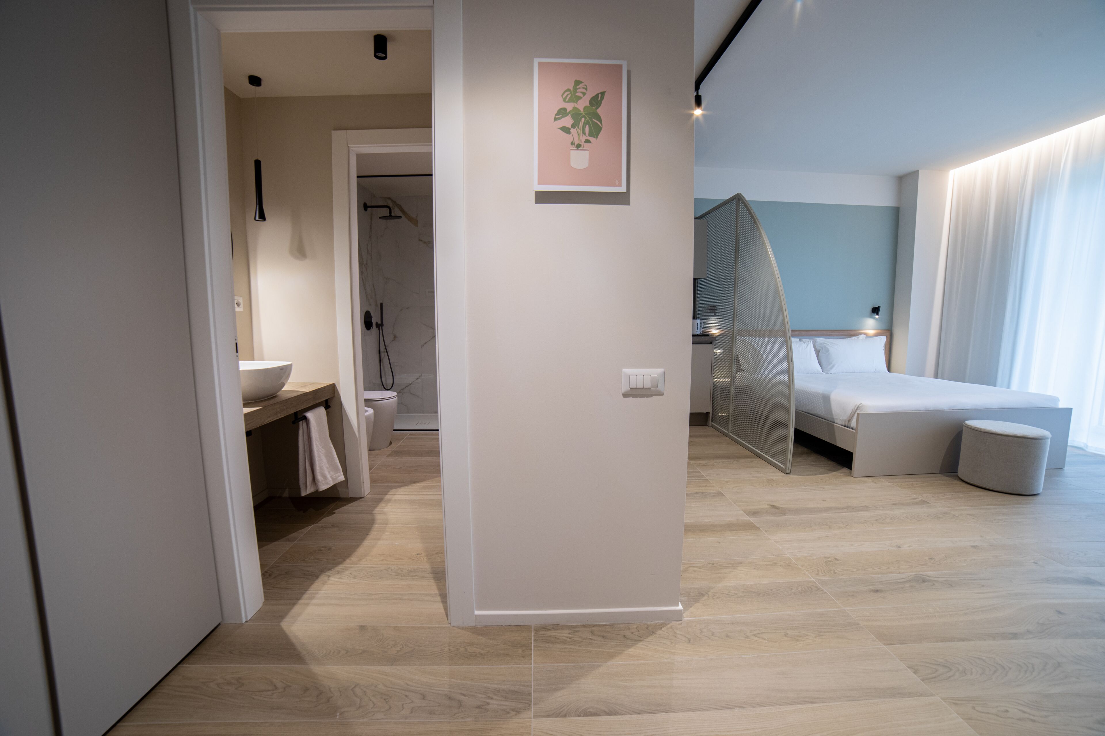 A modern hotel room with a wooden floor, a bed with a canopy, a bedside table, and a bathroom with a sink and towel rack.