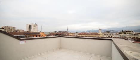 A rooftop terrace with a tiled floor, a view of surrounding buildings, and a cloudy sky.