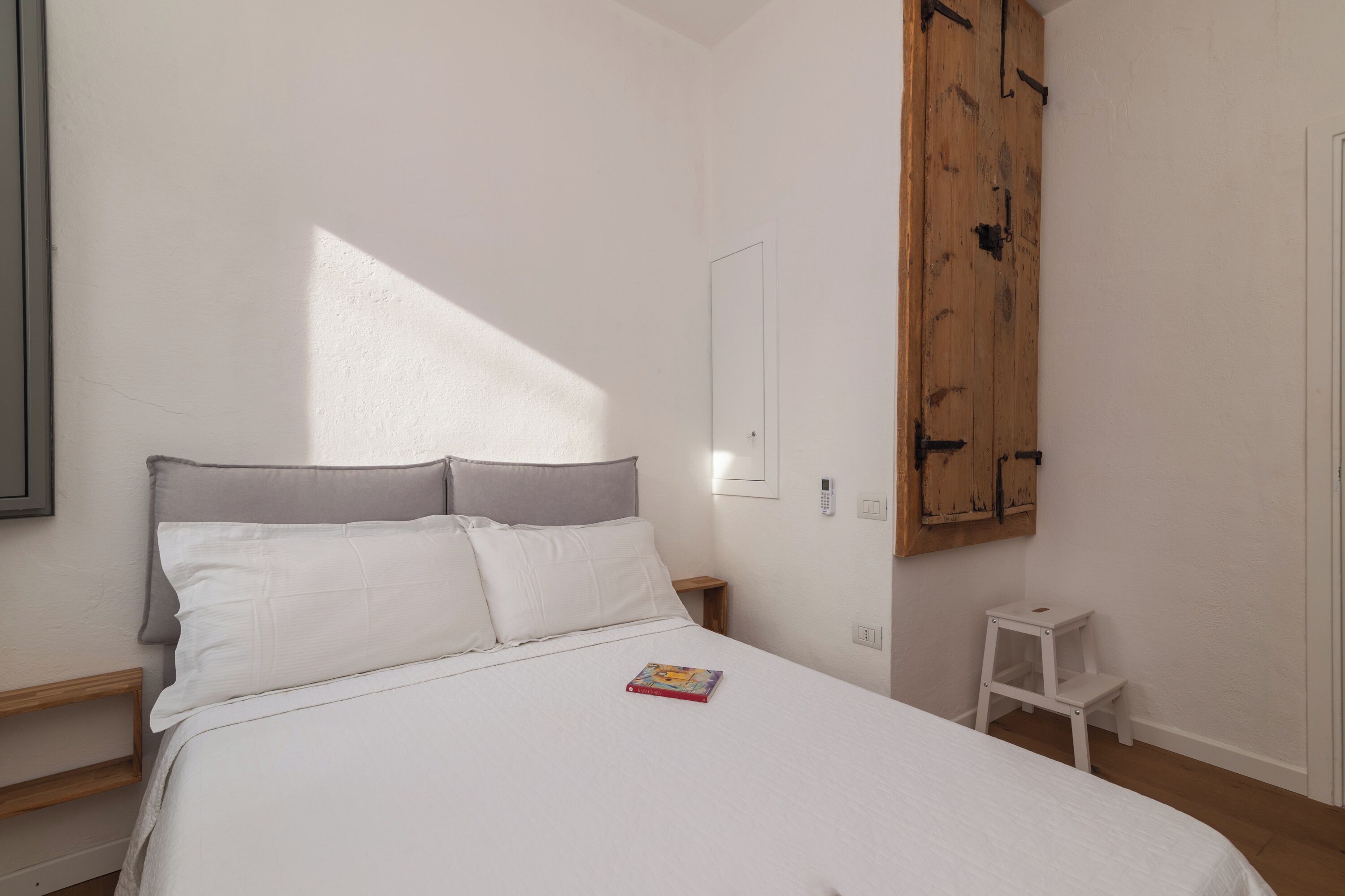 Une chambre à coucher avec un lit, une petite table de chevet blanche, une armoire en bois et une fenêtre qui projette une ombre.
