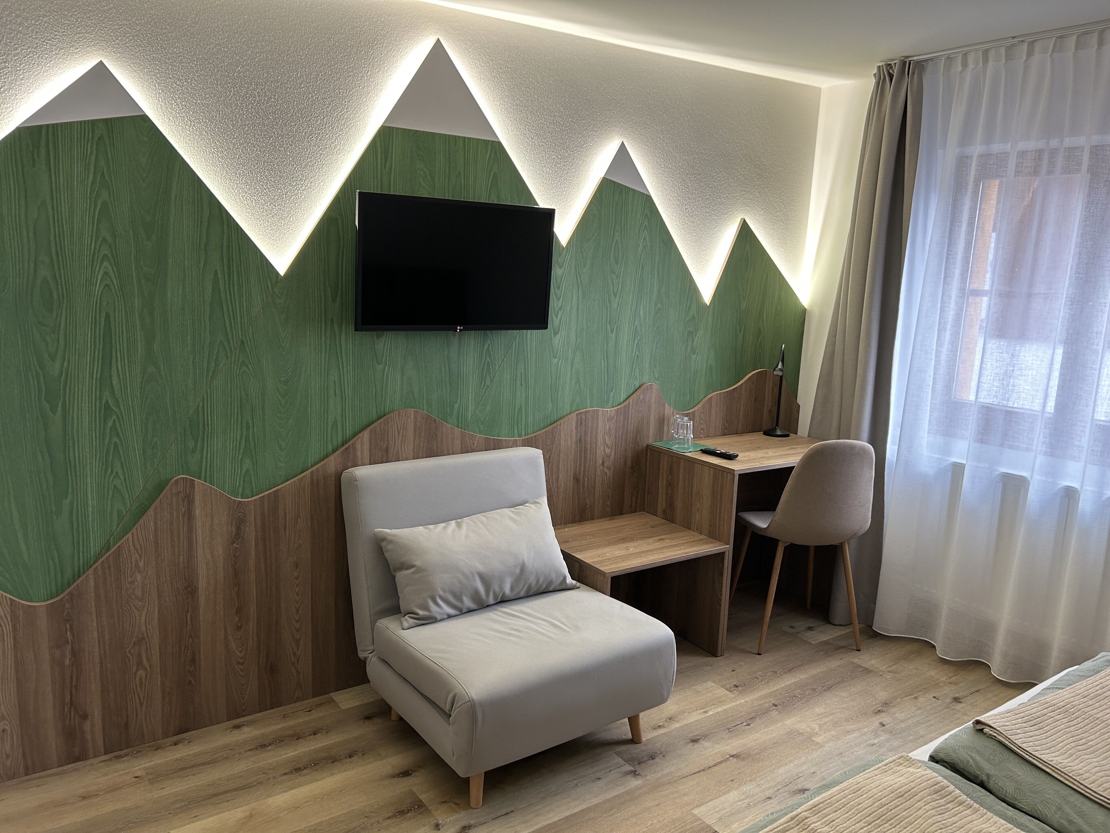 Una habitación de hotel moderna con una cama, un escritorio y un televisor montado en una pared verde con paneles de madera.